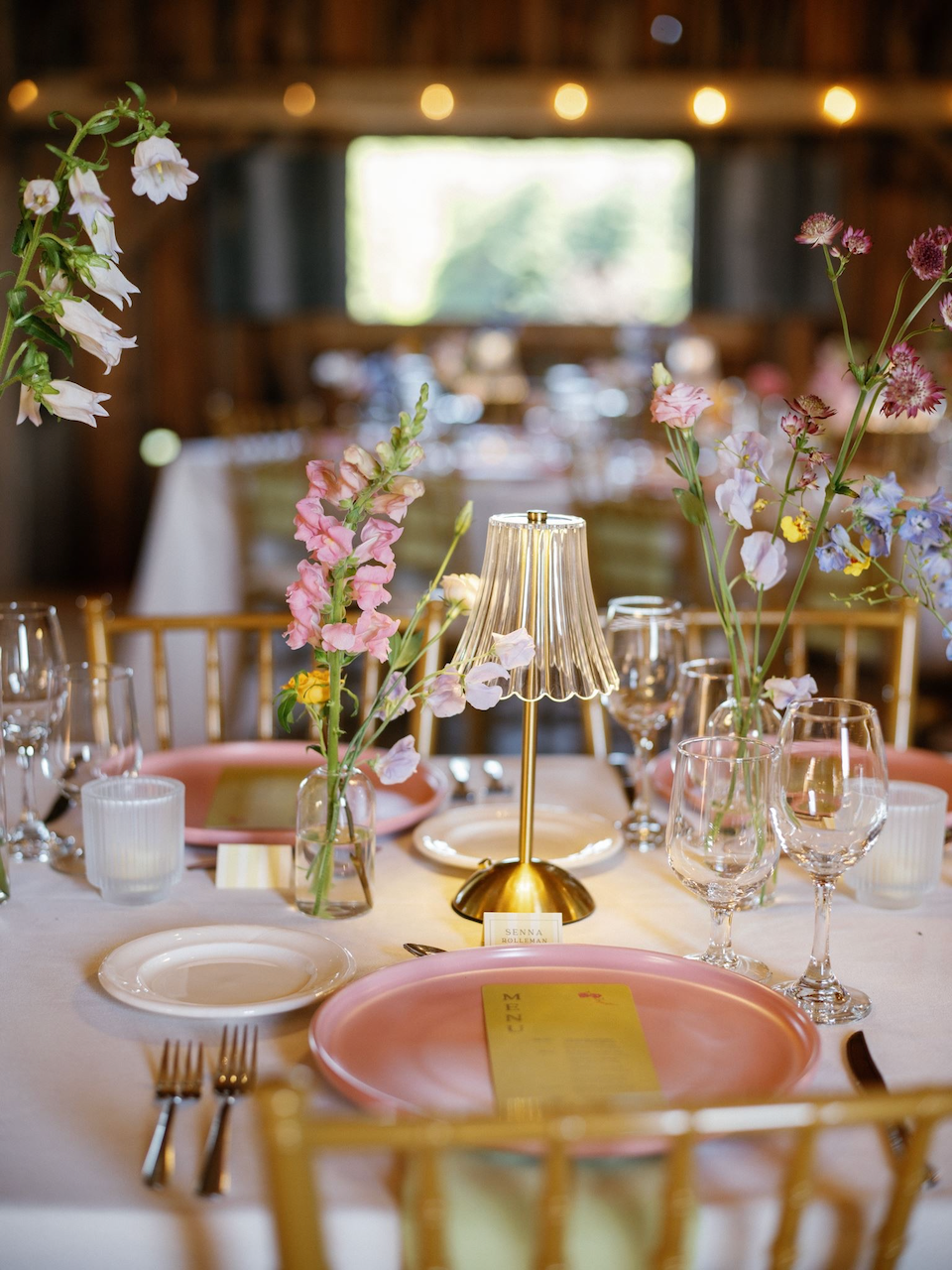A beautifully decorated table with pink plates, glassware, a golden lamp, and floral centerpieces in a cozy, rustic setting with string lights.