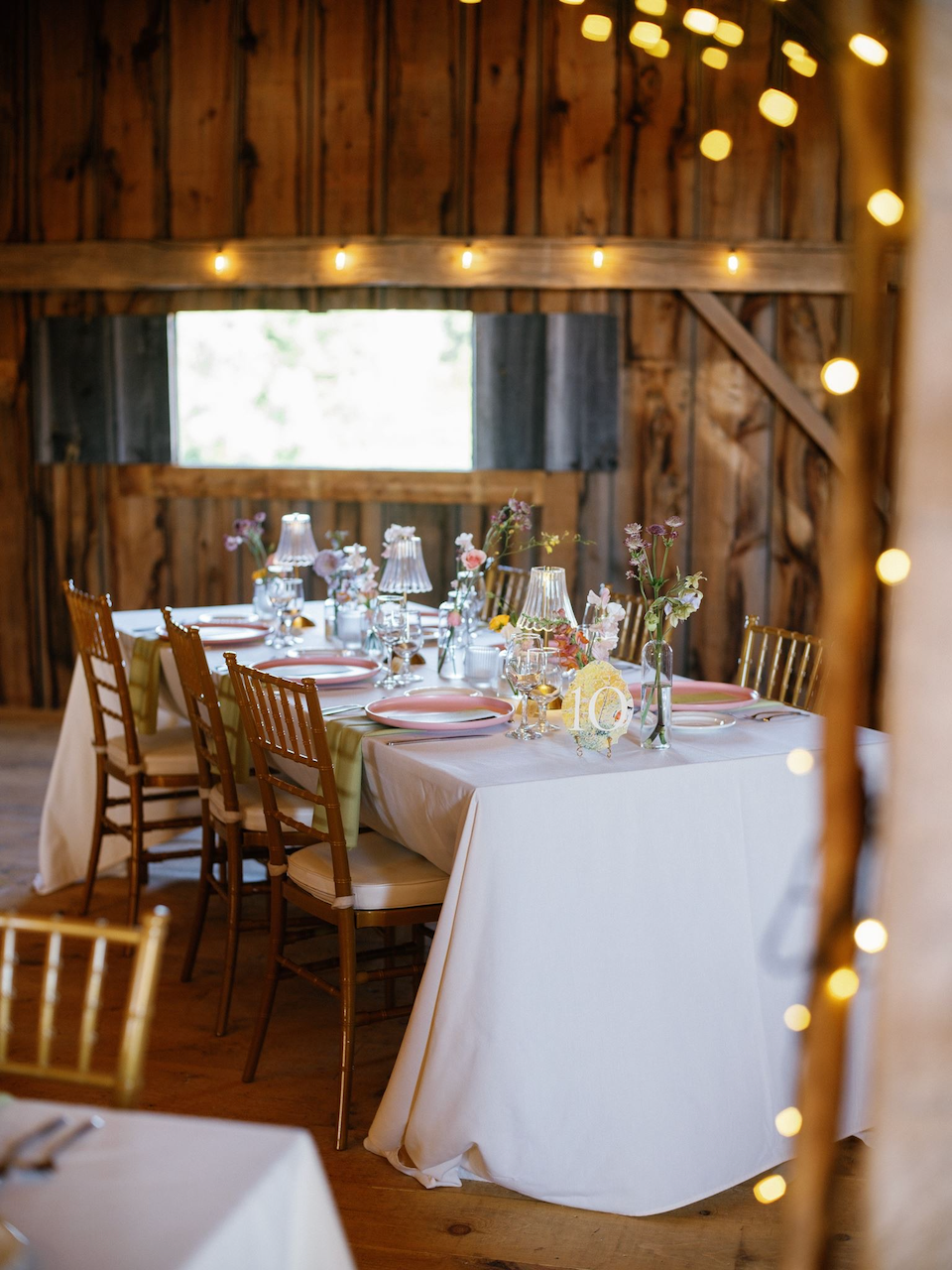 A rustic wedding reception table decorated with pink petals, glass vases with pink and purple flowers, small table lamps with pleated shades, pink plates, and glasses, set in a barn with wooden walls and fairy lights.