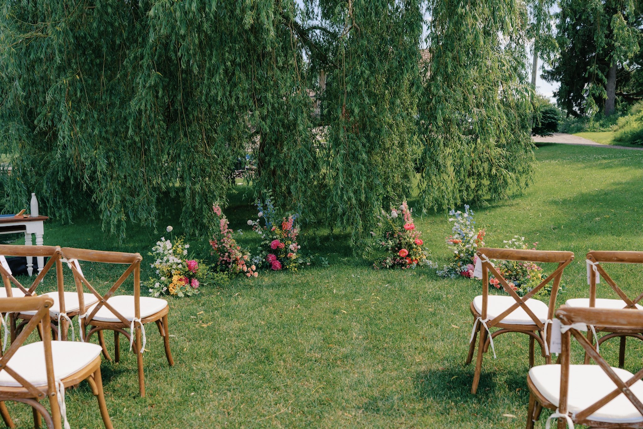 Ceremony chairs with white cushions set up outdoors for a wedding with a garden arrangement of colorful flowers and a large willow tree in the background.