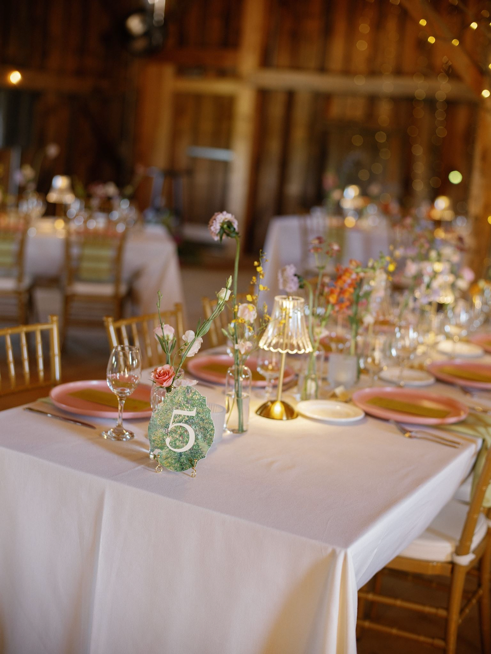 A decorated banquet table in a rustic wooden venue with pink and gold accents, floral centerpieces, table number 5, and soft lighting.