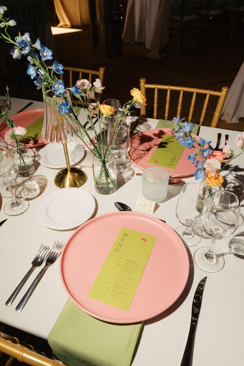 A table set for a meal with pink plates, green napkins, utensils, wine glasses, and a floral centerpiece with blue, pink, and yellow flowers. There is a gold lamp with a pink shade on the table.