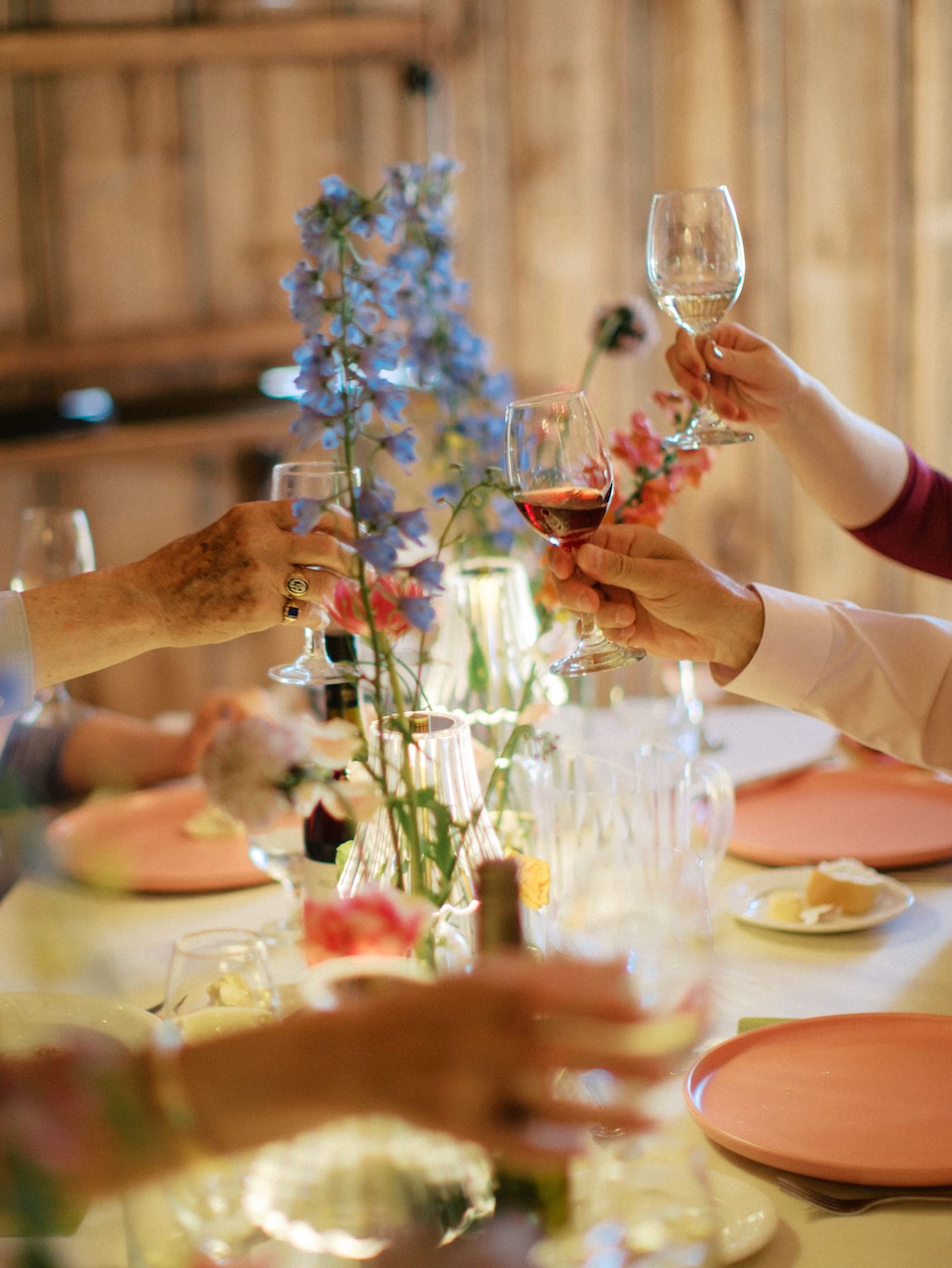 People raising glasses of red and white wine in a toast at a dinner party with floral centerpieces on the table.