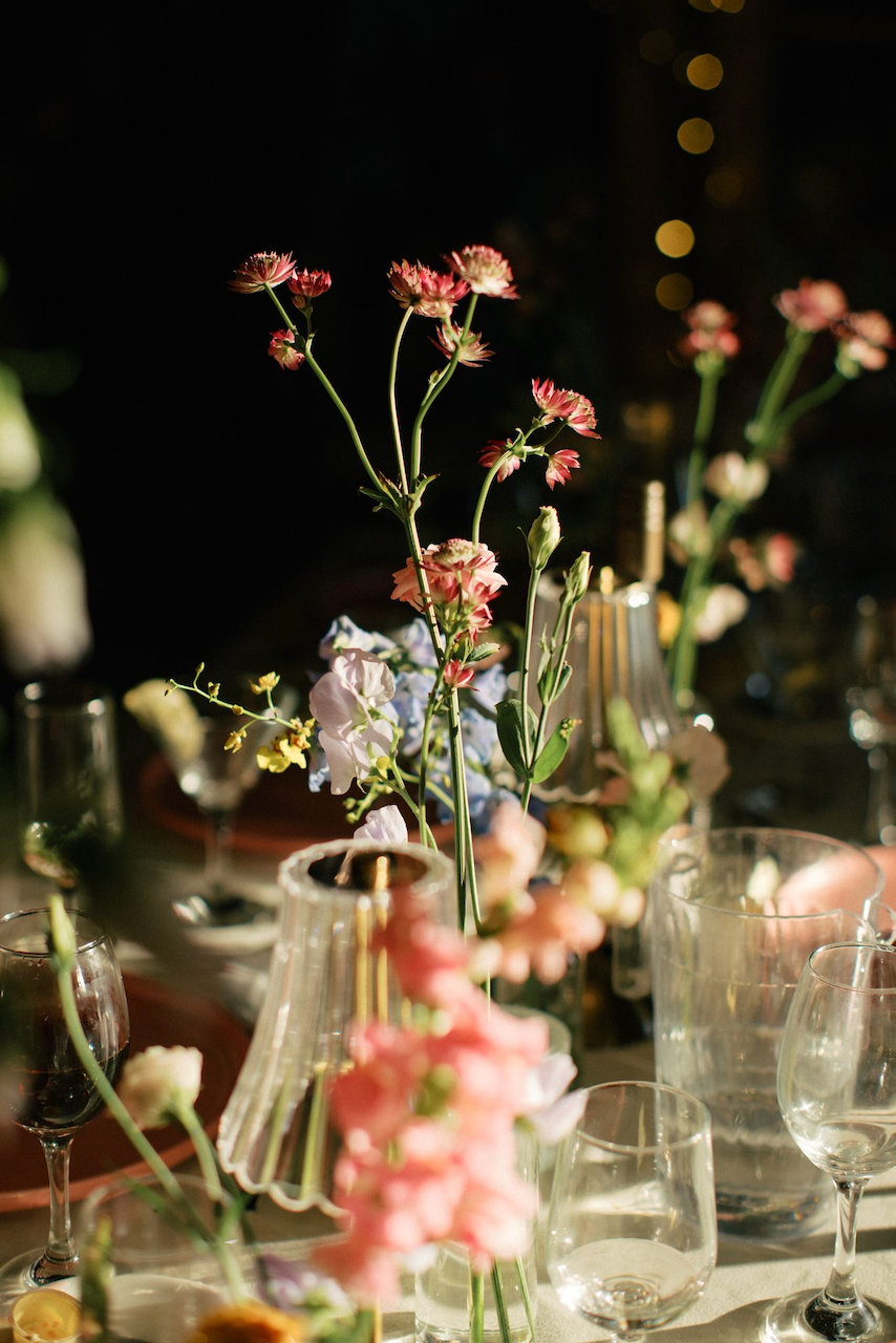 A close-up of tall pink and white flowers in a vase on a table set for a dinner or event, with other flowers and glassware around.