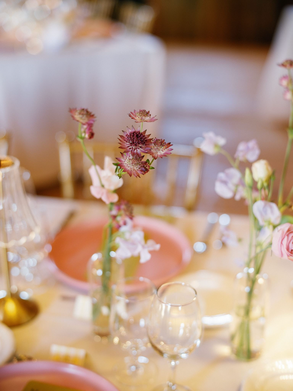 Flowers in small vases on a banquet table with pink plates, wine glasses, and gold accents at an elegant event.