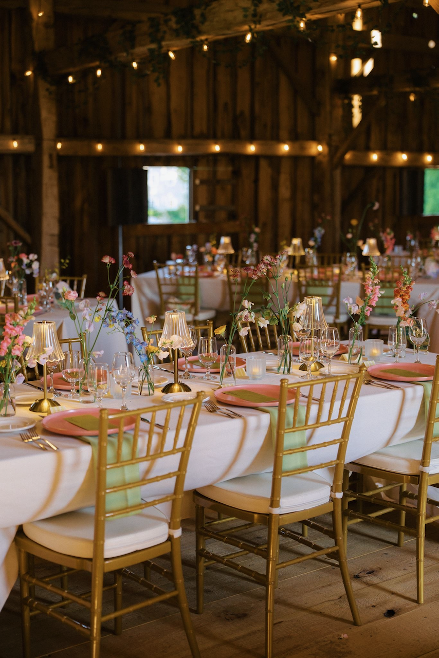 A decorated rustic barn interior set up for a wedding or event with a long white-clothed table, pink plates, gold chairs, floral centerpieces, and warm string lights overhead.