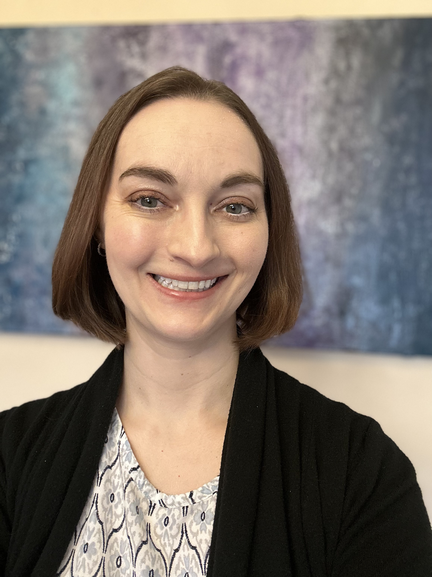 A woman smiling, with shoulder-length brown hair, green eyes, and wearing a black jacket over a patterned blouse, standing in front of an abstract purple and blue background.