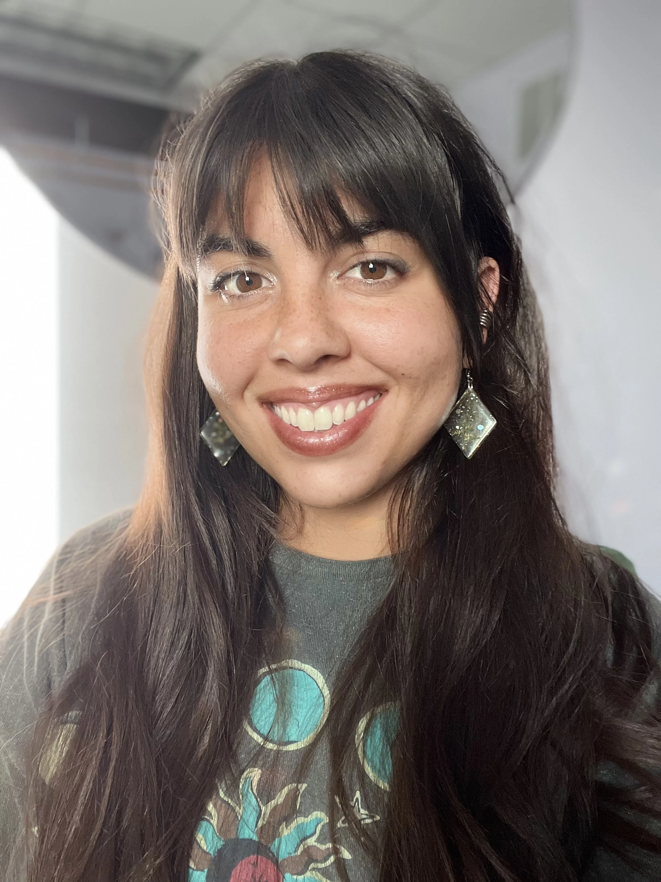 Close-up of a woman with long brown hair, wearing earrings and a graphic T-shirt, smiling at the camera.