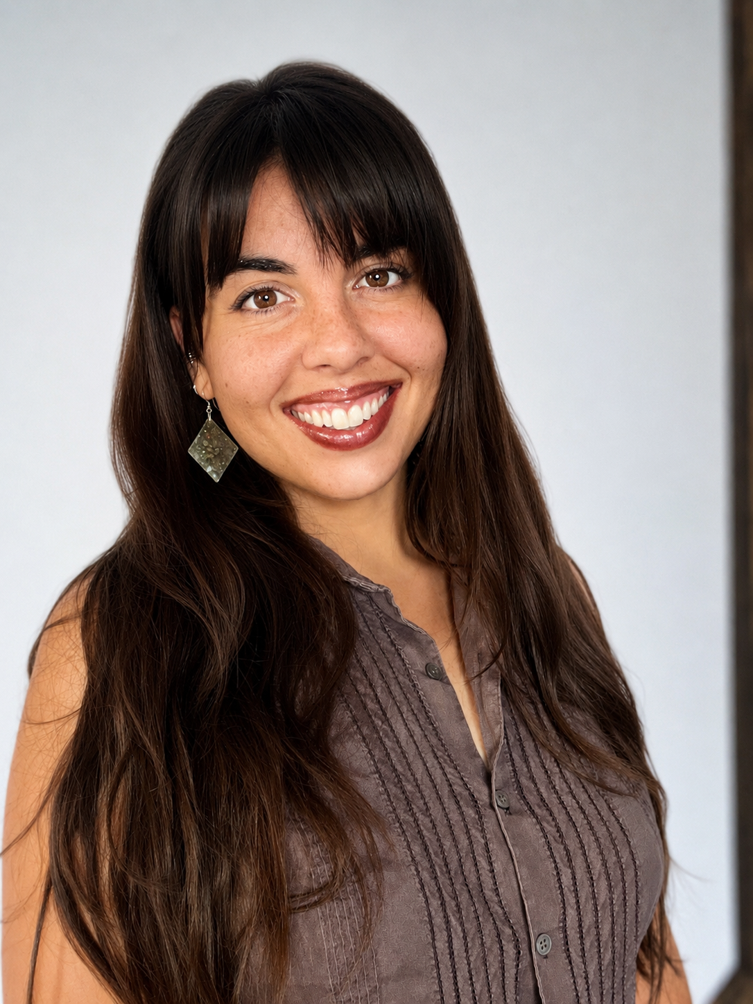 A woman with long dark hair, wearing a sleeveless button-up shirt and earrings, smiling against a plain background.