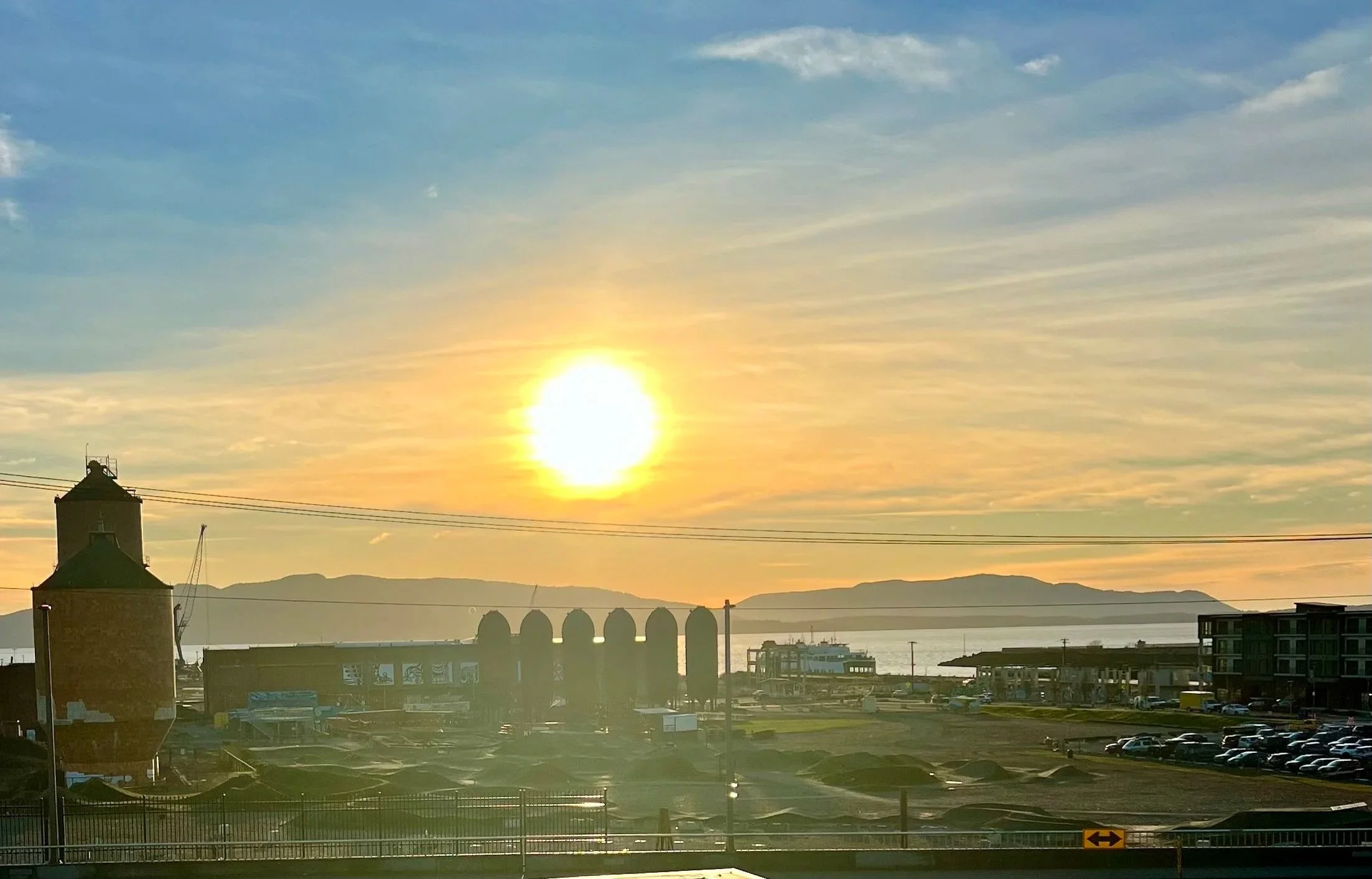 Sunset over a city with rooftops, a body of water, mountains in the background, and partly cloudy blue sky.