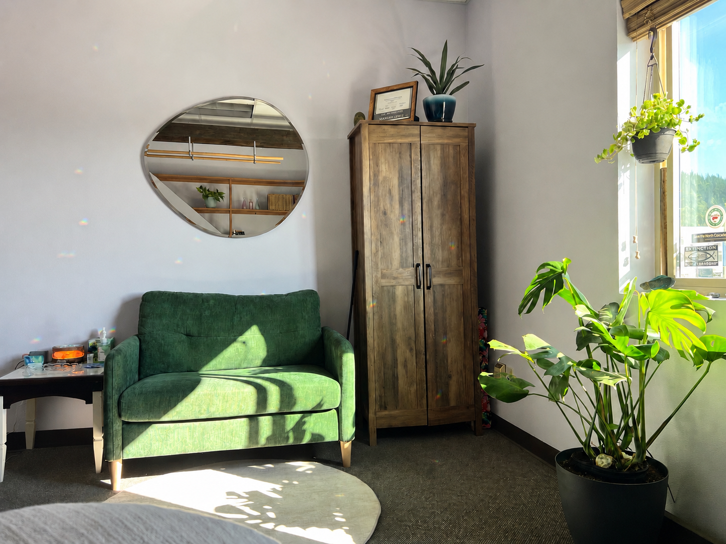 A cozy corner of a living room with a green upholstered loveseat, a small black side table with toiletries, a wooden wardrobe, a large potted plant, and a window with natural sunlight and hanging plants.