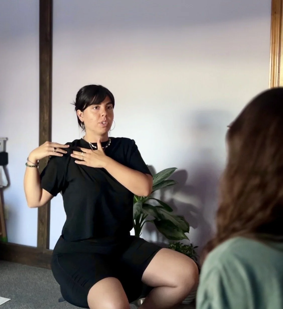 Woman sitting and speaking in a casual indoor setting, gesturing with her hands and wearing a black outfit, with another person partially visible in the foreground.