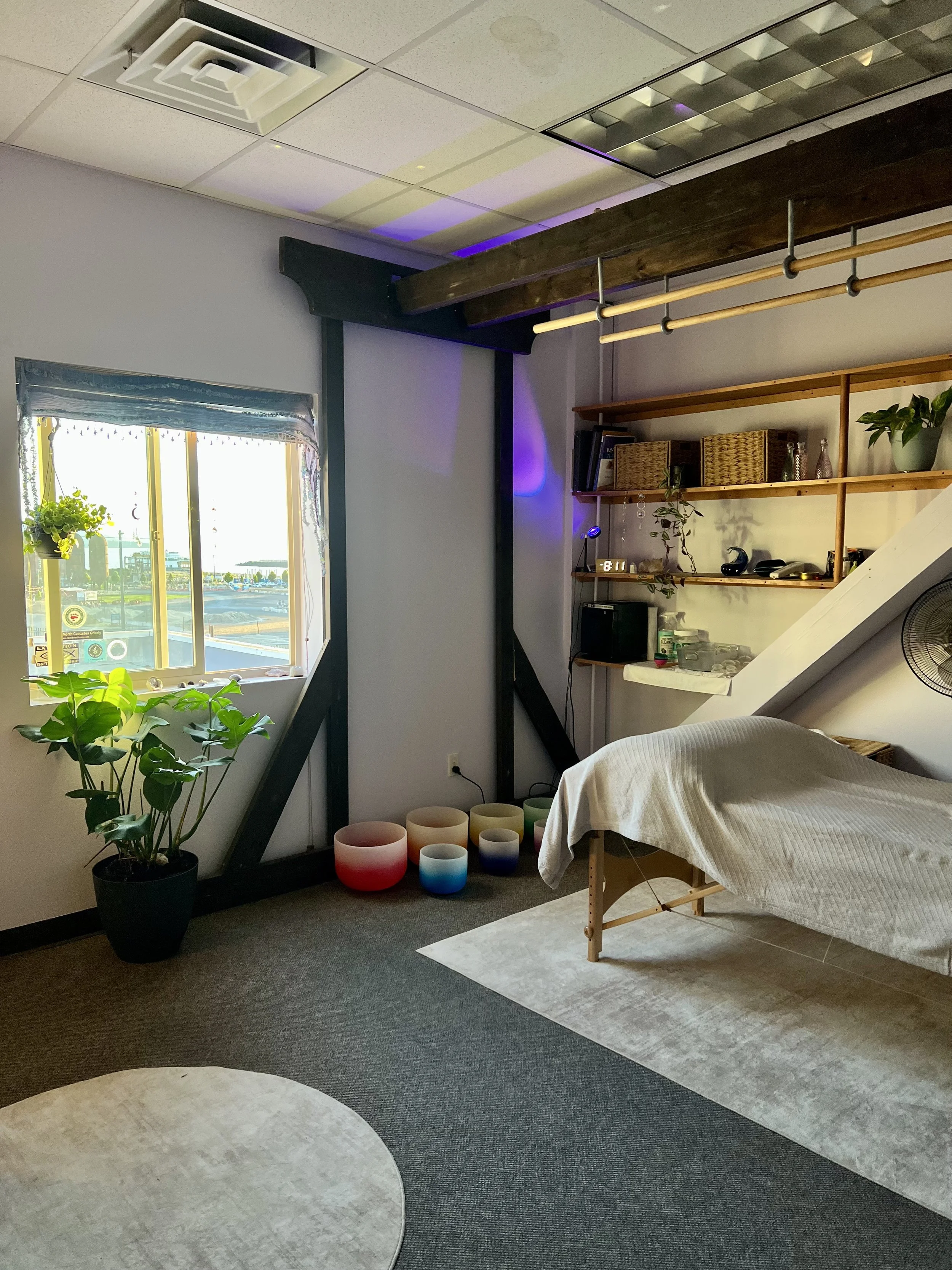 A cozy room with a large window that has a view of an outdoor area, a potted plant near the window, wobbly stained glass bowls in front of the window, a light-colored blanket on a wooden bed frame, a wooden shelf with various decorations and books, a hanging wooden rack, and a ceiling with air vents and lights.