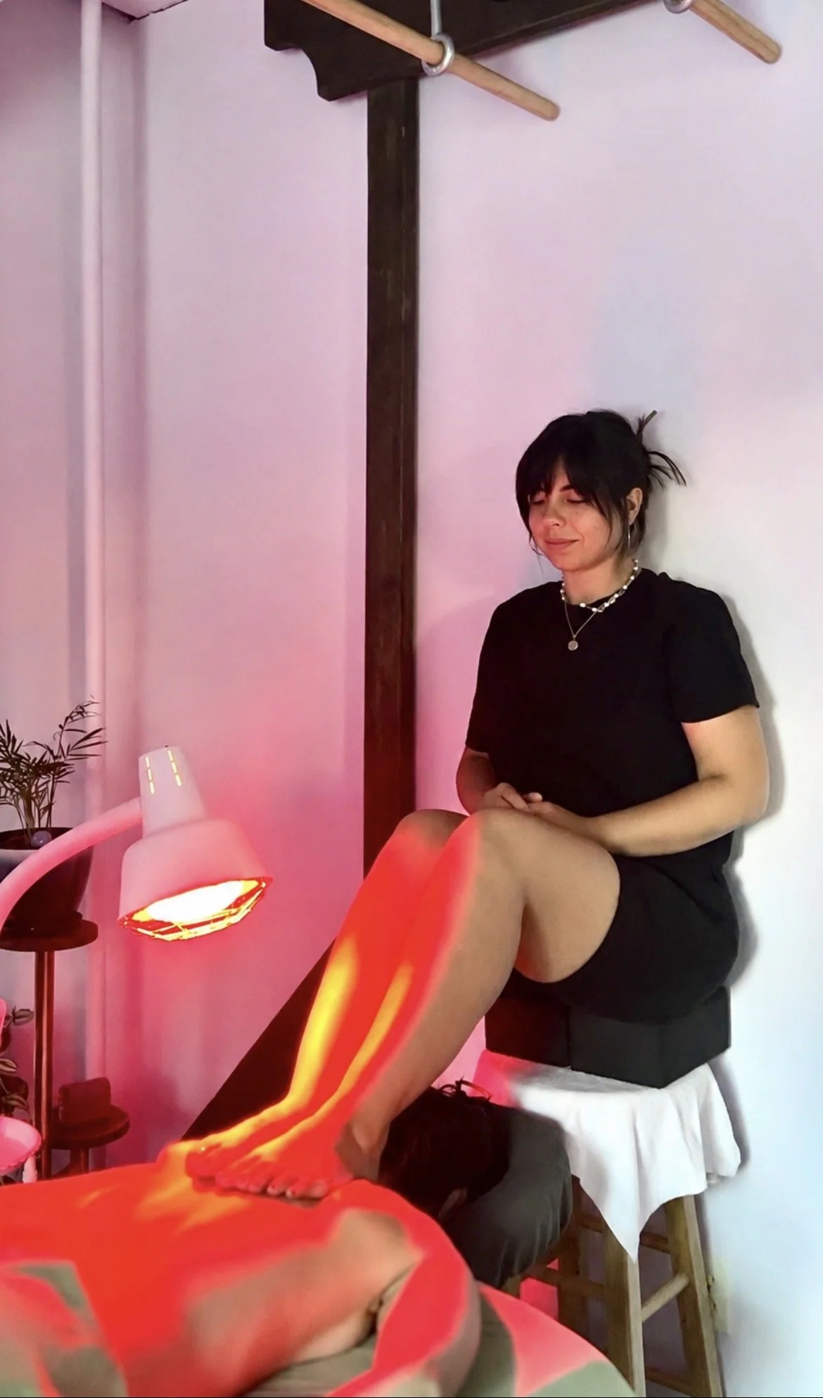 A woman with short black hair and necklaces sitting with her eyes closed, receiving a foot massage from a person lying on a massage table. The room has pink and purple lighting, a small lamp, and a potted plant nearby.