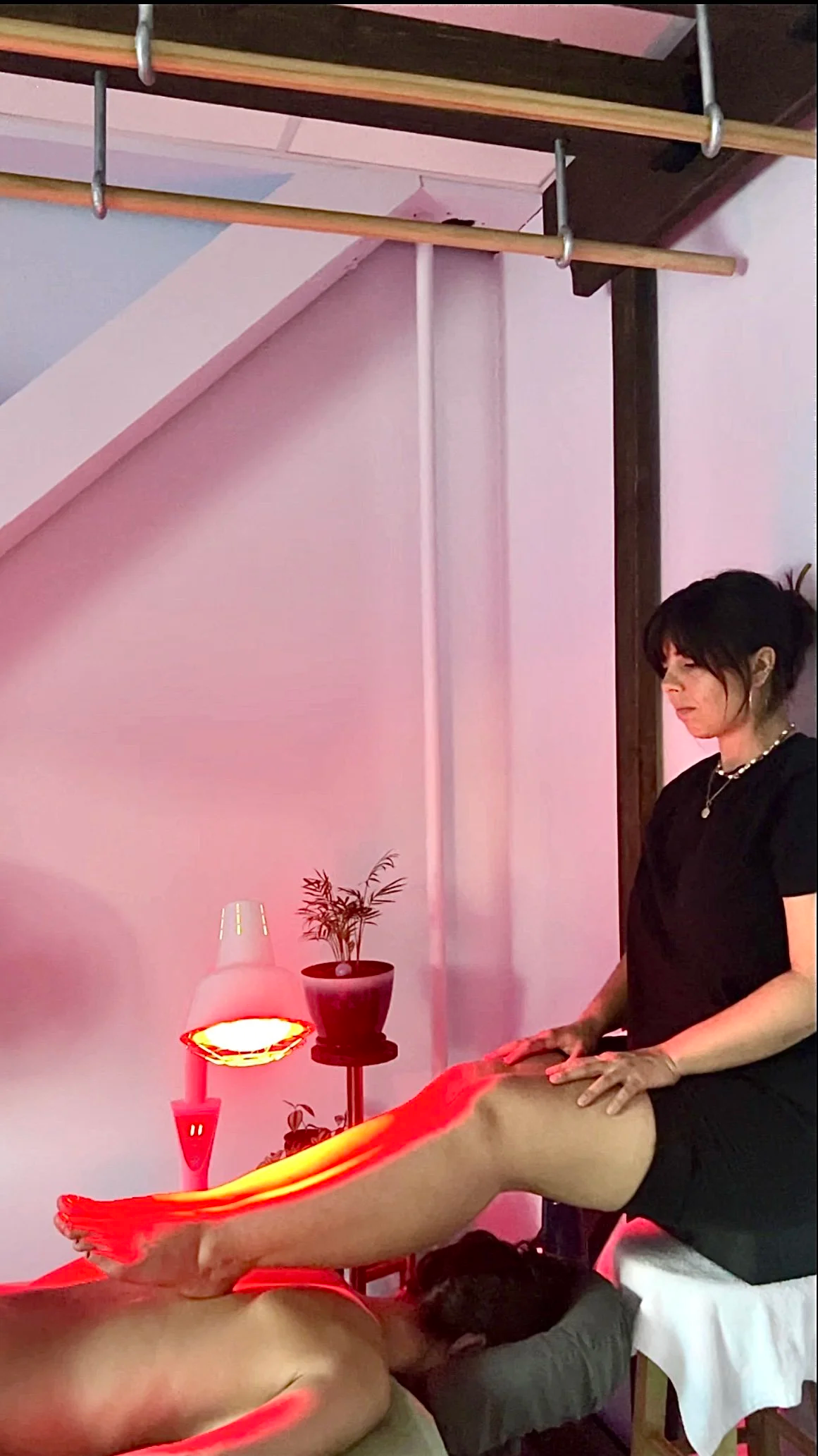 A woman giving a massage to a person lying face down on a massage table, with a pink and red ambient lighting and a small bedside table with a lamp and plant.