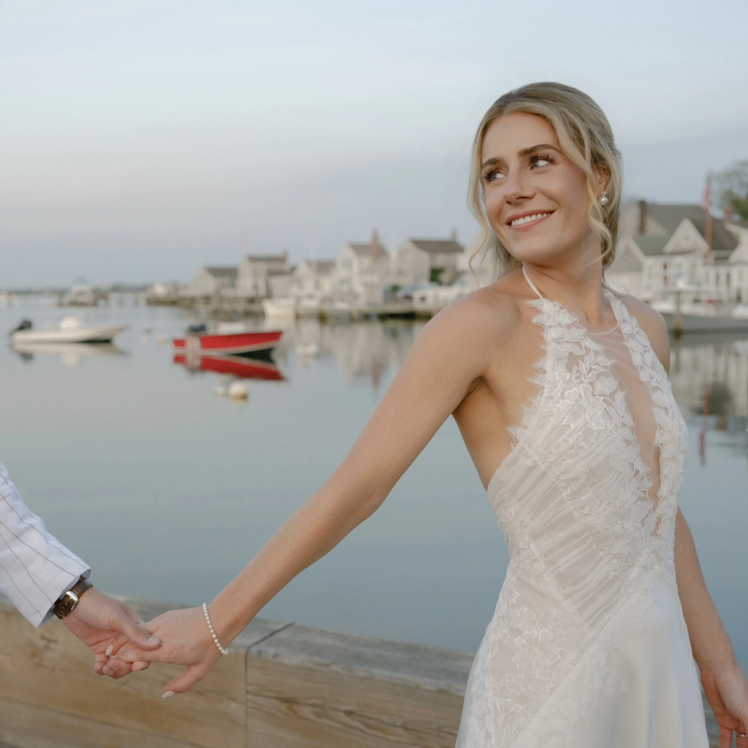 A woman in a white lace dress smiling and holding hands with a man in a white shirt, standing near a harbor with boats and houses in the background.