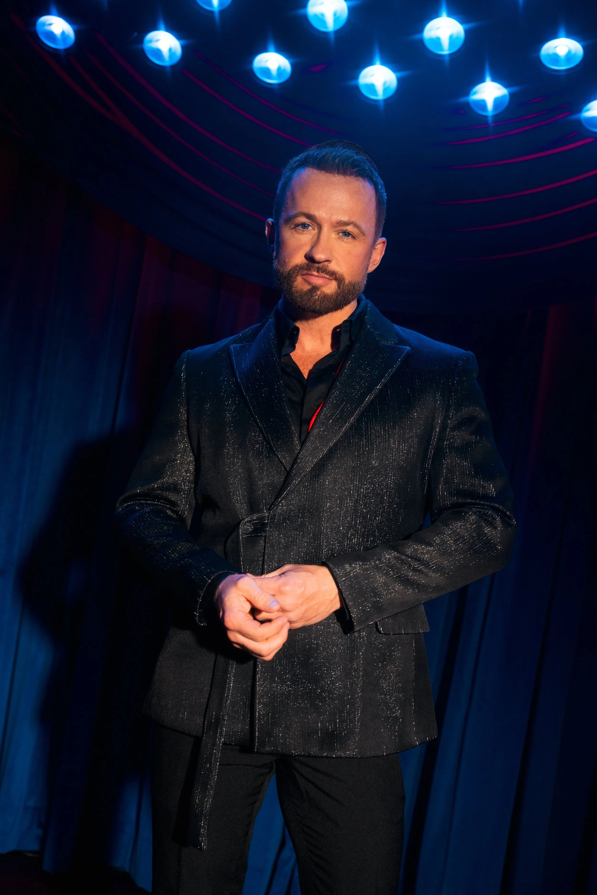 Marty Thomas - a member of the trio Vocal group Triple Threat. A man with a beard and short dark hair, wearing a shiny black suit, standing in front of a dark background with hanging blue lights.