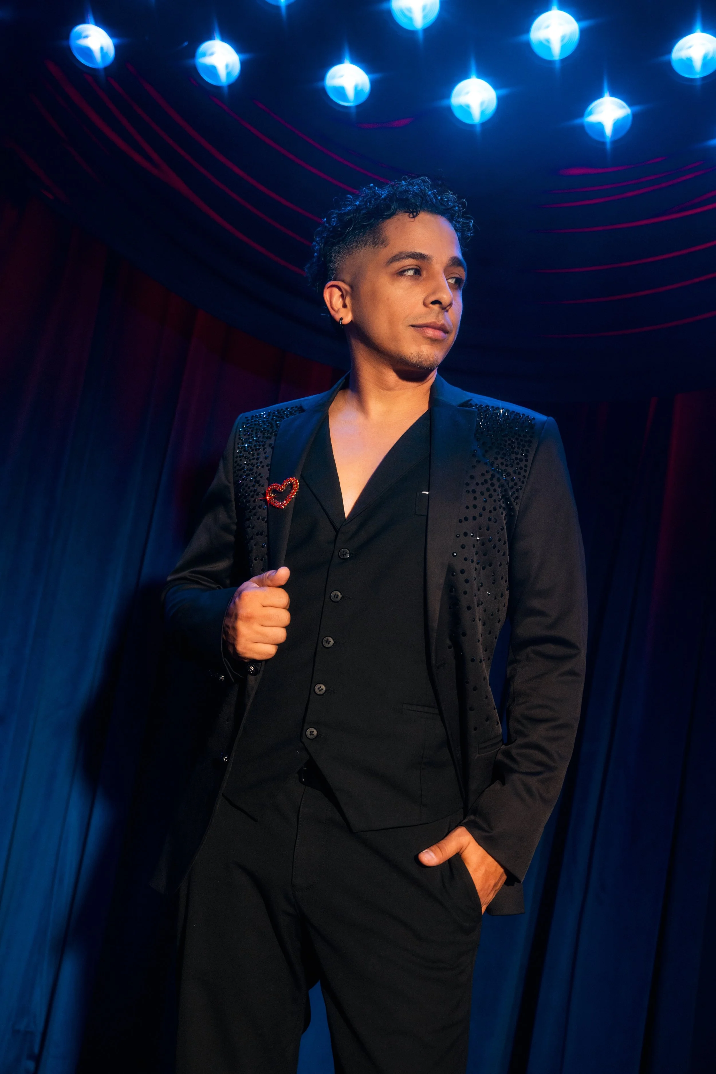 Omar Cardona - a member of the trio Vocal group Triple Threat. A man with short curly hair, wearing a black suit  stands in a dark room with blue lighting and red curved lines on the ceiling, looking to his right with one hand on his hip.