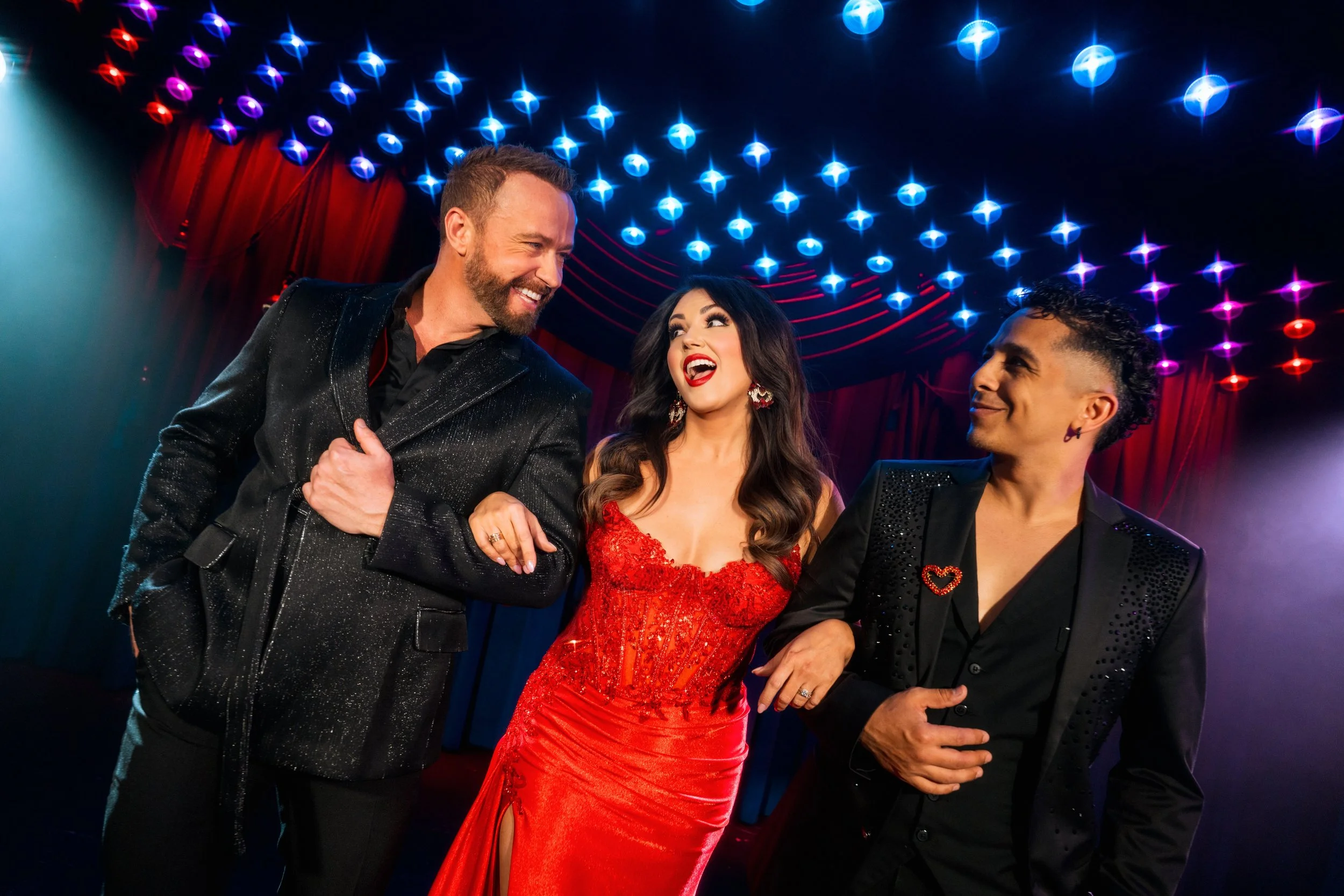 Triple Threat (trio vocal group) consisting of Marty Thomas, Rachel Potter, and Omar Cardona. Three people dressed in evening attire, smiling and talking, under colorful stage lights with red curtains in the background.