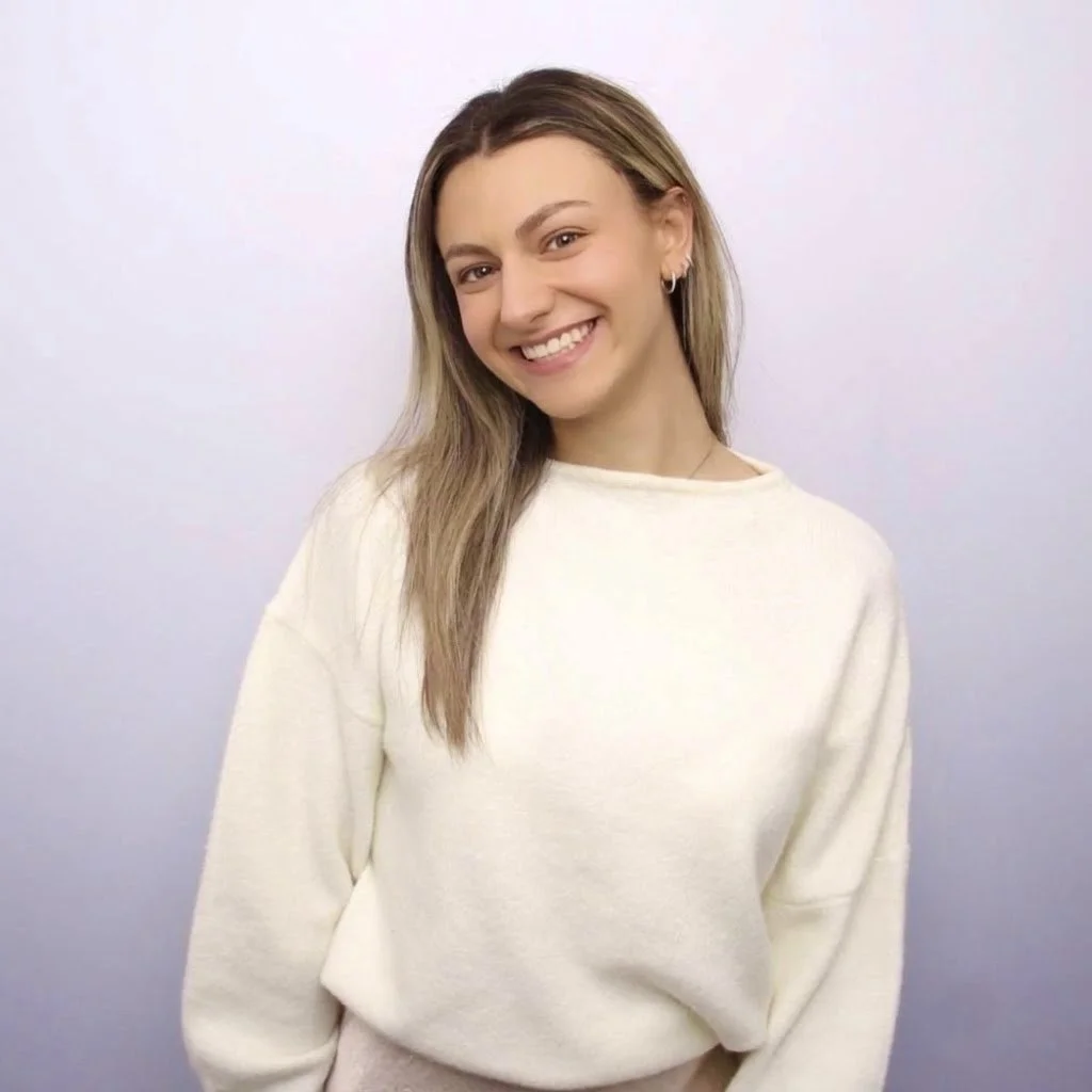 Smiling woman with long brown hair wearing a cream sweater against a pastel lavender background