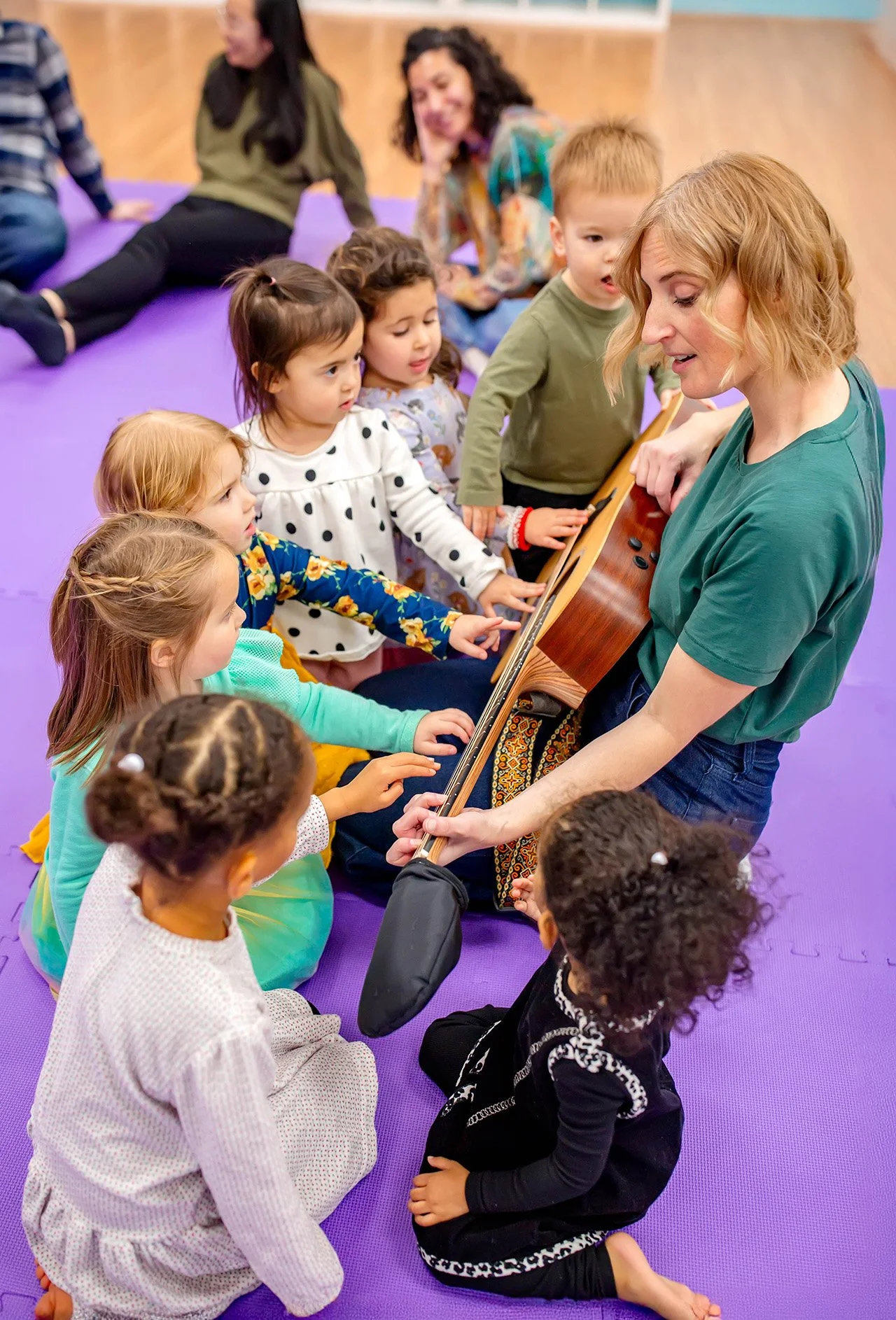 A group of young children sitting on a purple mat in a circle, engaged in a guitar lesson with a woman playing guitar. Two women are sitting in the background on the floor, one wearing a green jacket and the other with curly hair. The children are attentively reaching out to touch or strum the guitar.