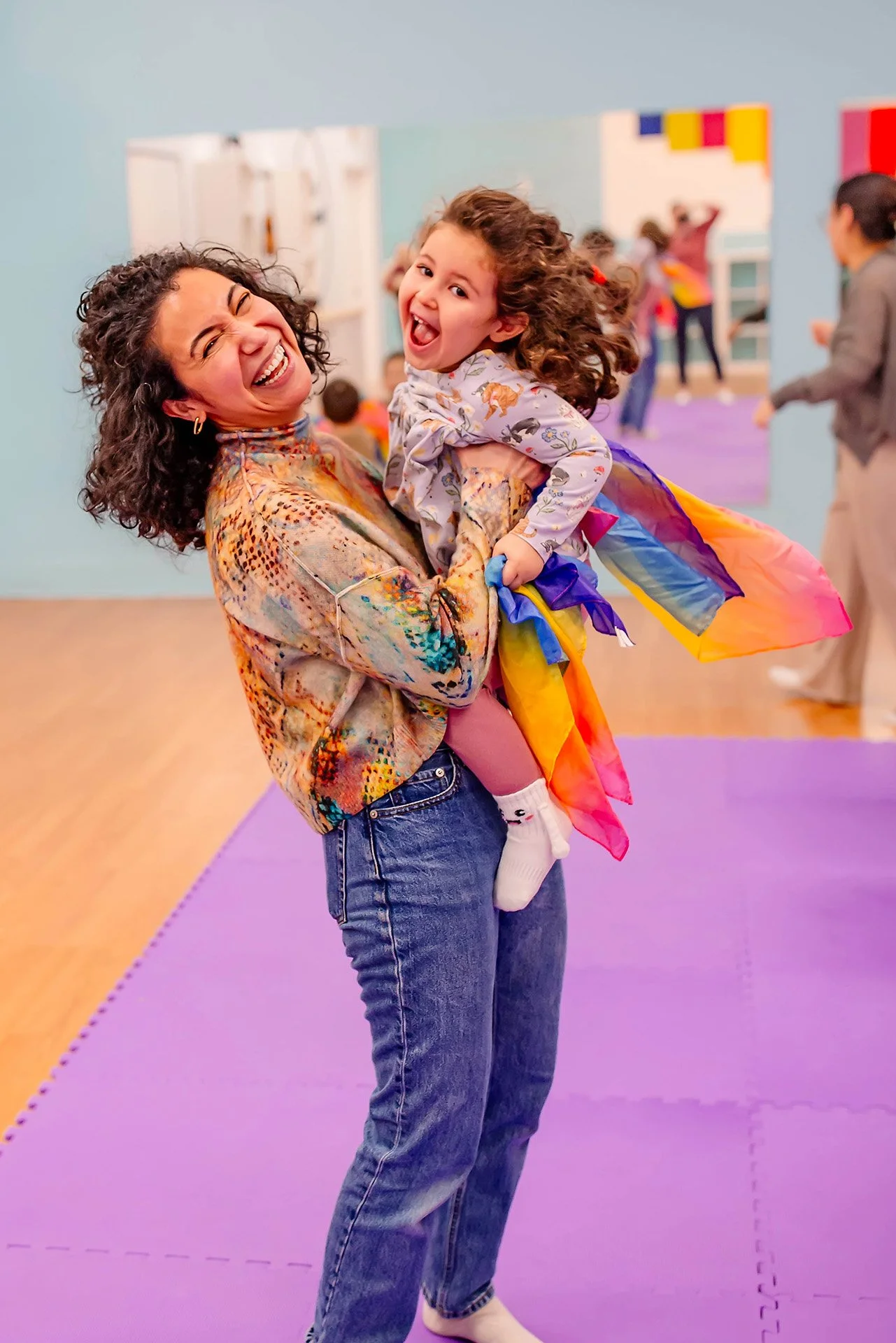 A woman with curly hair, wearing a colorful jacket and jeans, smiling and holding a young girl dressed as a rainbow-colored fairy with a rainbow tulle skirt in a playroom or gym with purple mats and other children in the background.