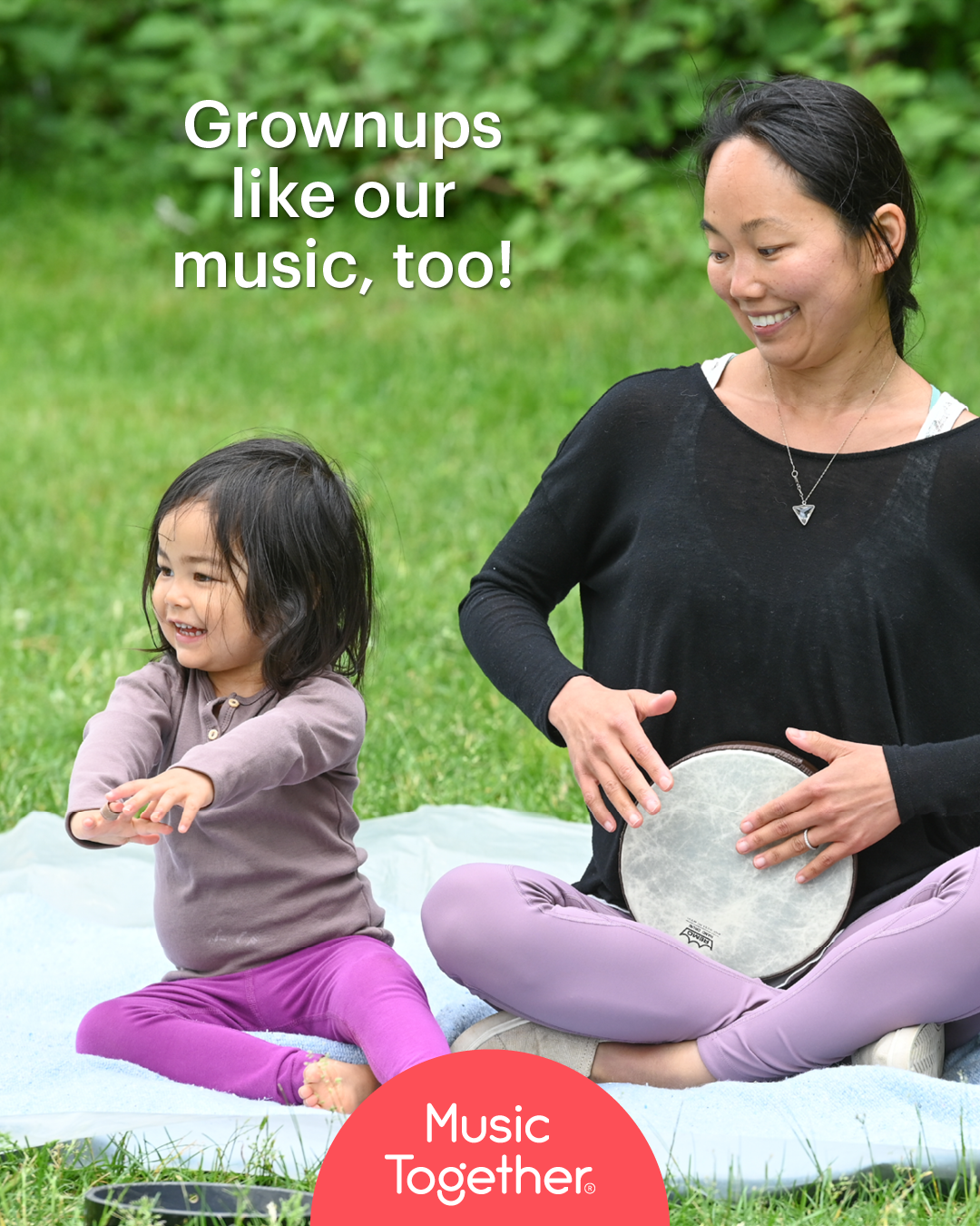 A woman and a young girl sitting on a blanket outdoors, playing musical instruments and smiling. The woman is holding a tambourine, and the girl appears to be clapping or playing along.