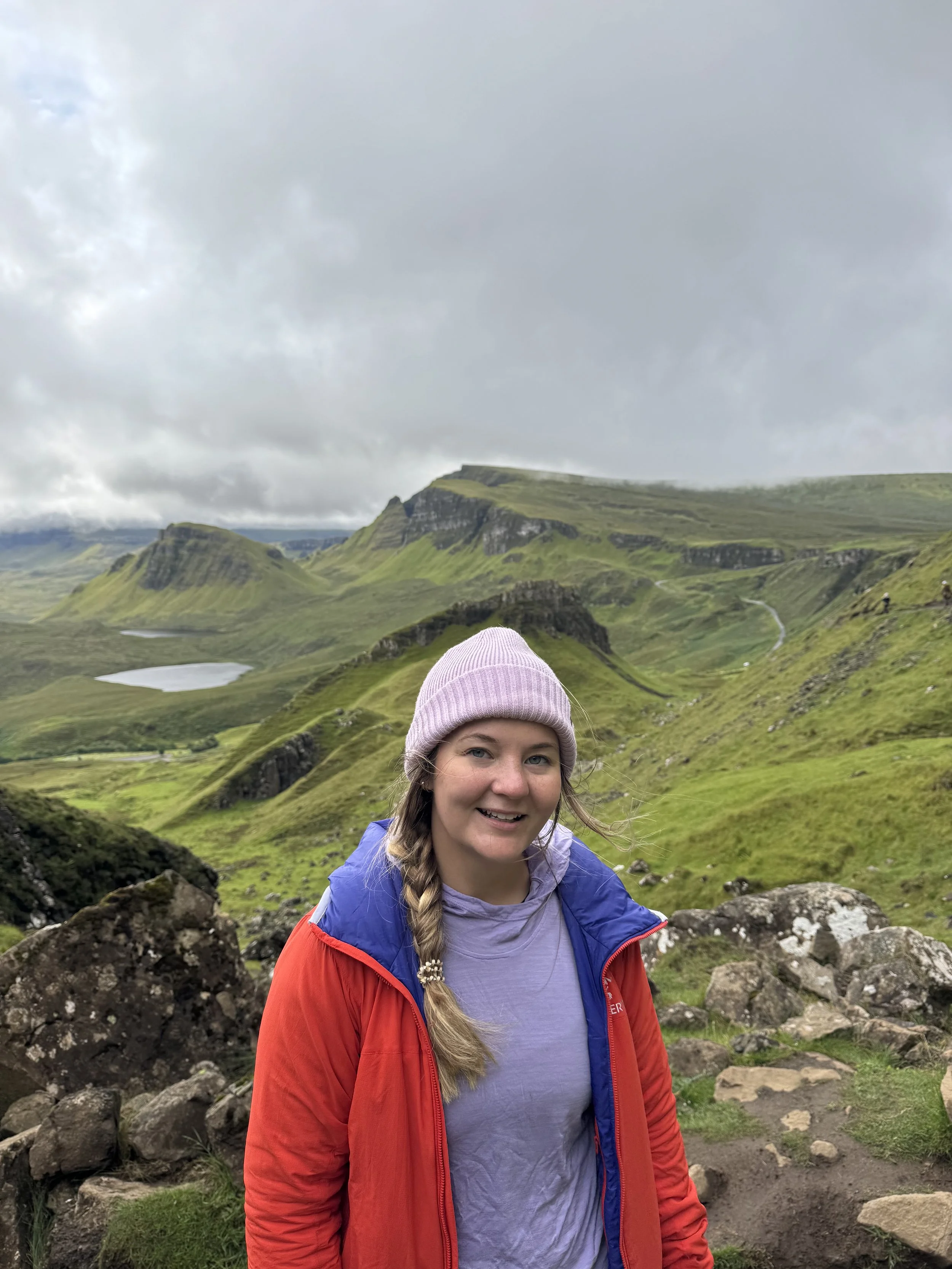A woman with blonde hair in a braid, wearing a pink beanie, purple shirt, and red jacket, smiling outdoors with green mountains and cloudy sky in the background.