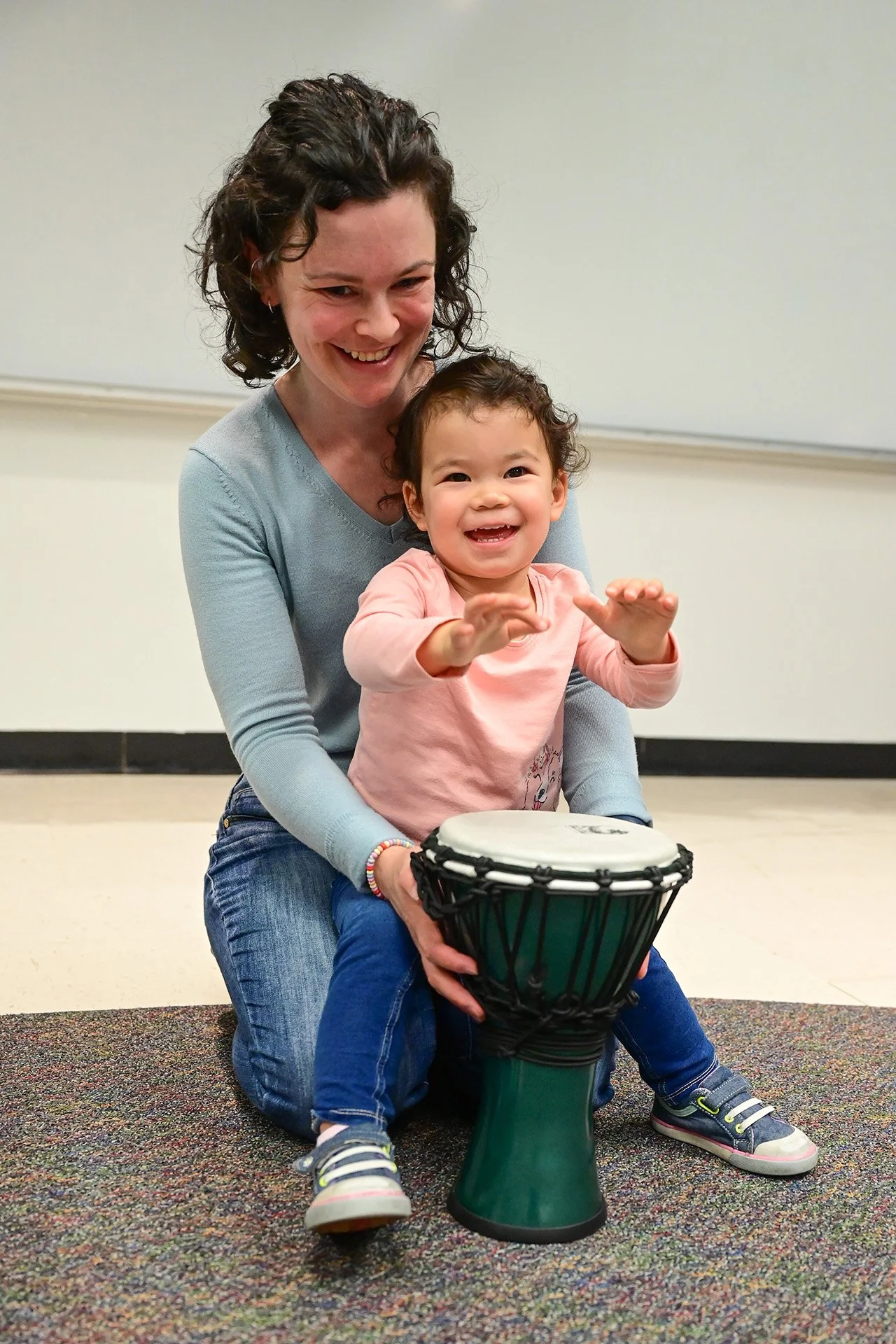 A woman and a young girl smiling and playing a drum together indoors, on a multicolored carpet.