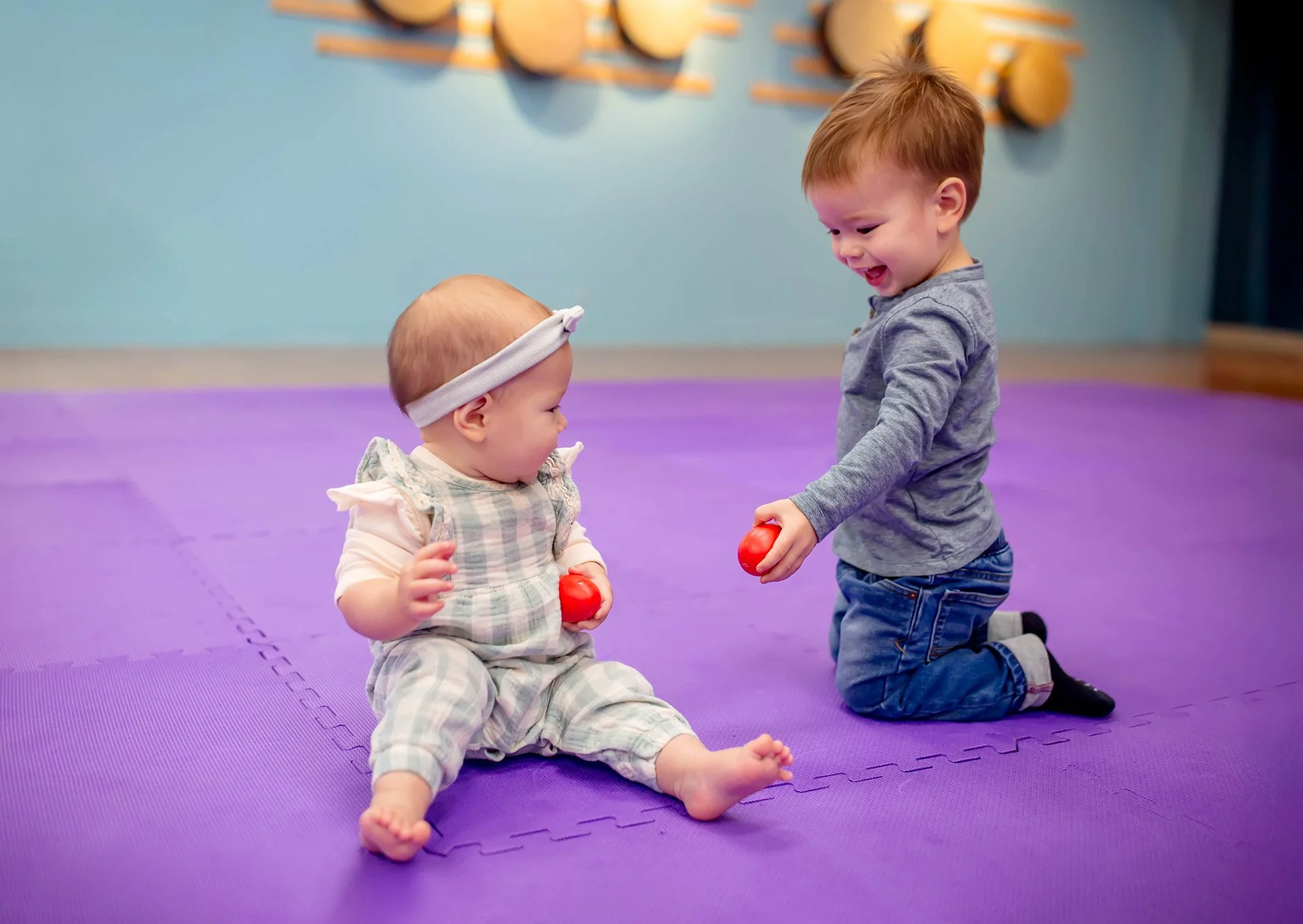 Two young children, a seated baby girl and a kneeling boy, playing with red balls on a purple mat in a playroom with drum decorations on the wall.