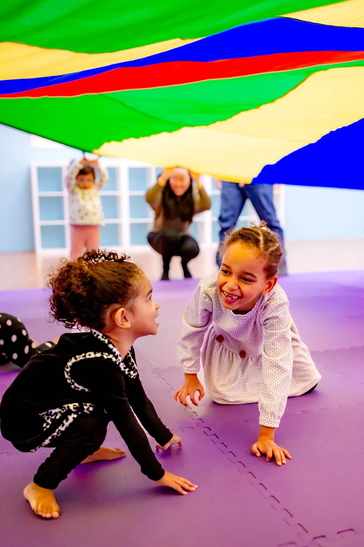 Two young children with curly hair on purple mat engaging in conversation, with other children and colorful parachute overhead in a playroom.