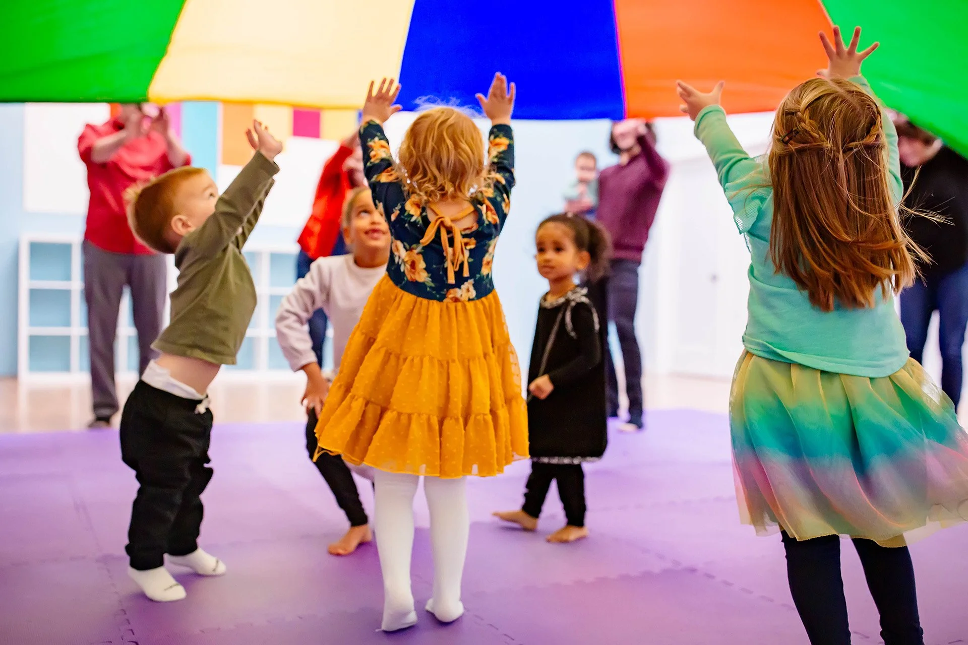 Children participating in a group activity with a colorful parachute indoors, some children are reaching up to hold the parachute, while others are smiling and watching.