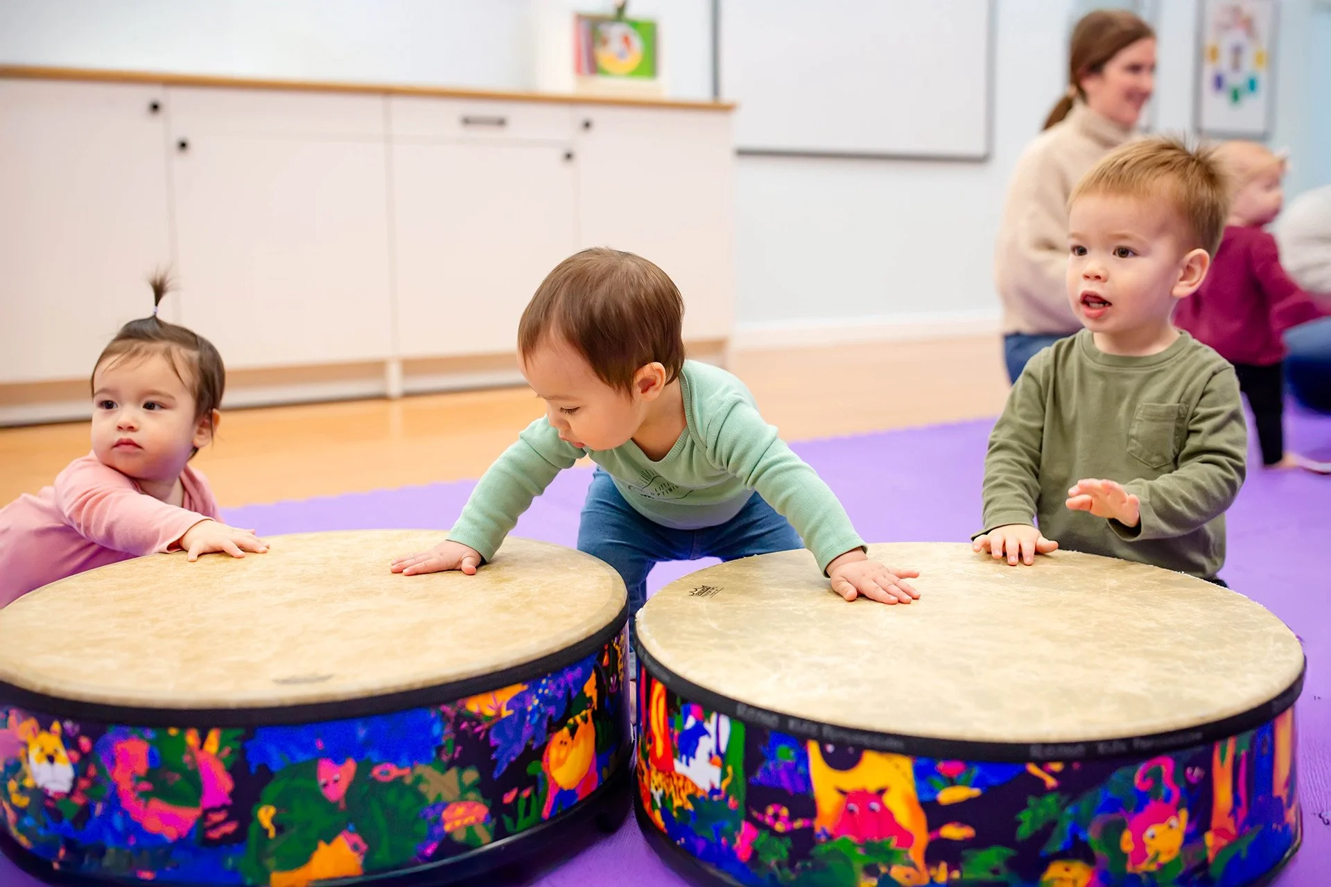 Children playing musical drums in a classroom with a teacher in the background.