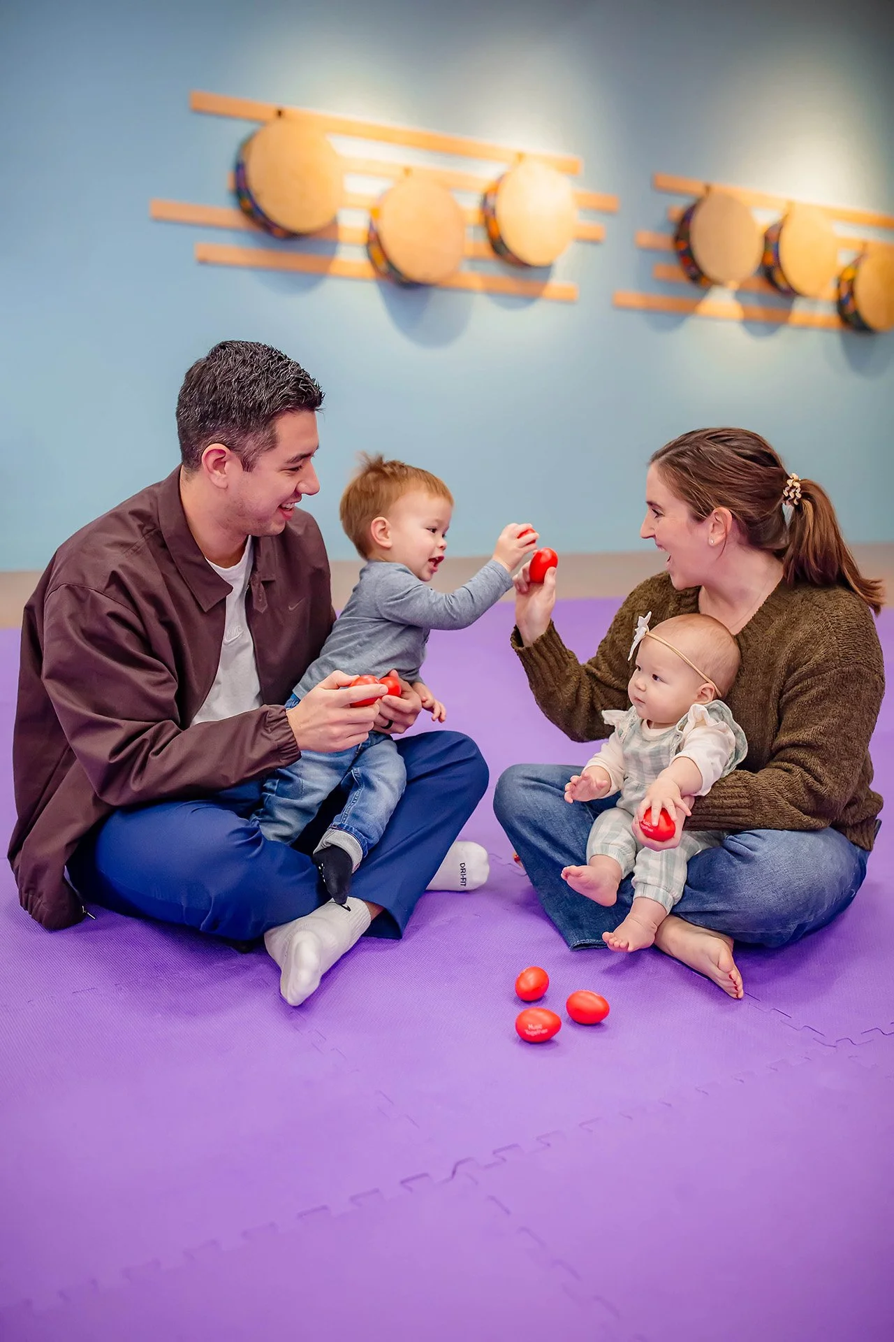 A family of four sitting on a purple exercise mat sharing tomatoes, with blue wall background and decorative wall art.