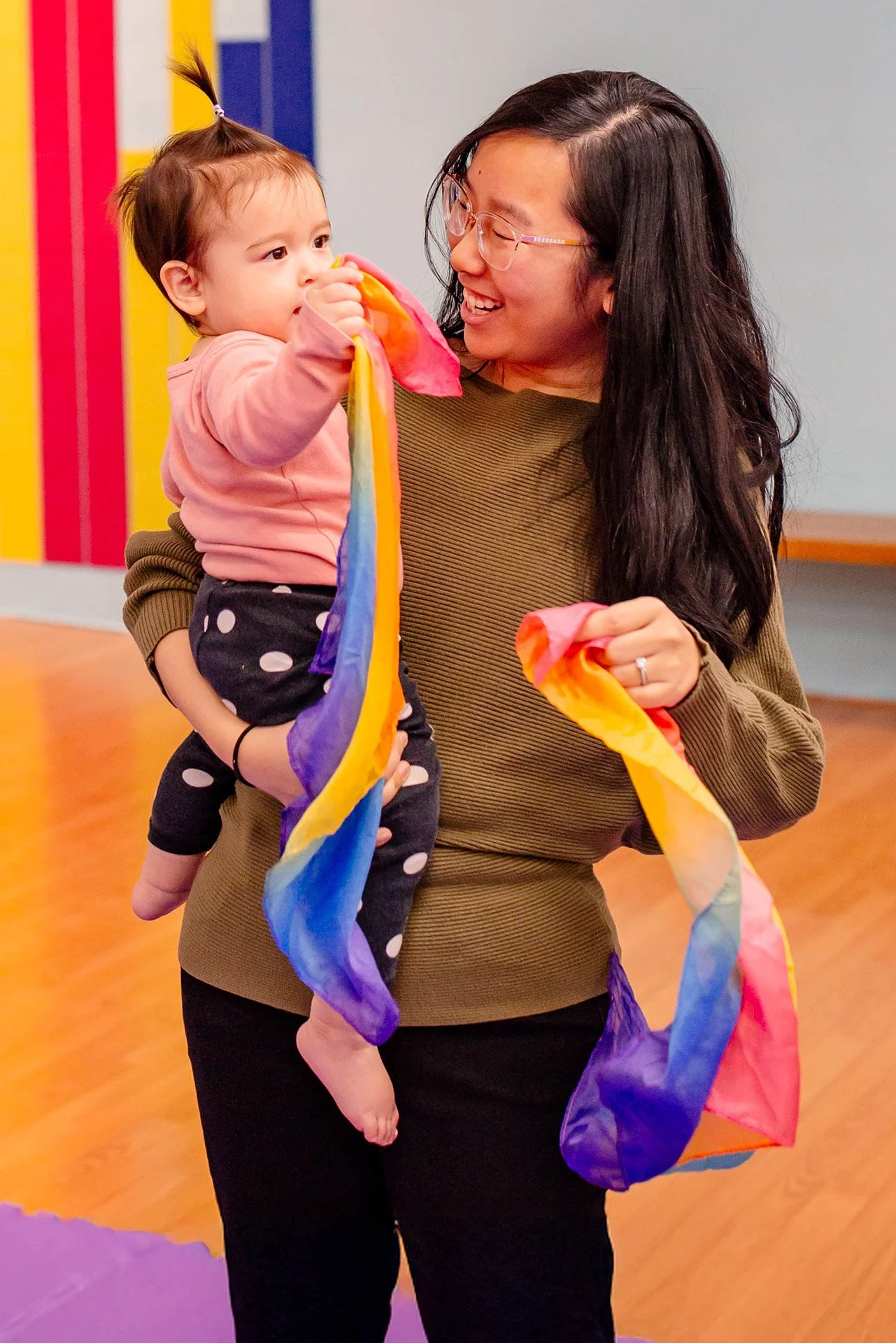 A woman holding a young girl in her arms while playing with colorful streamers in a playroom.