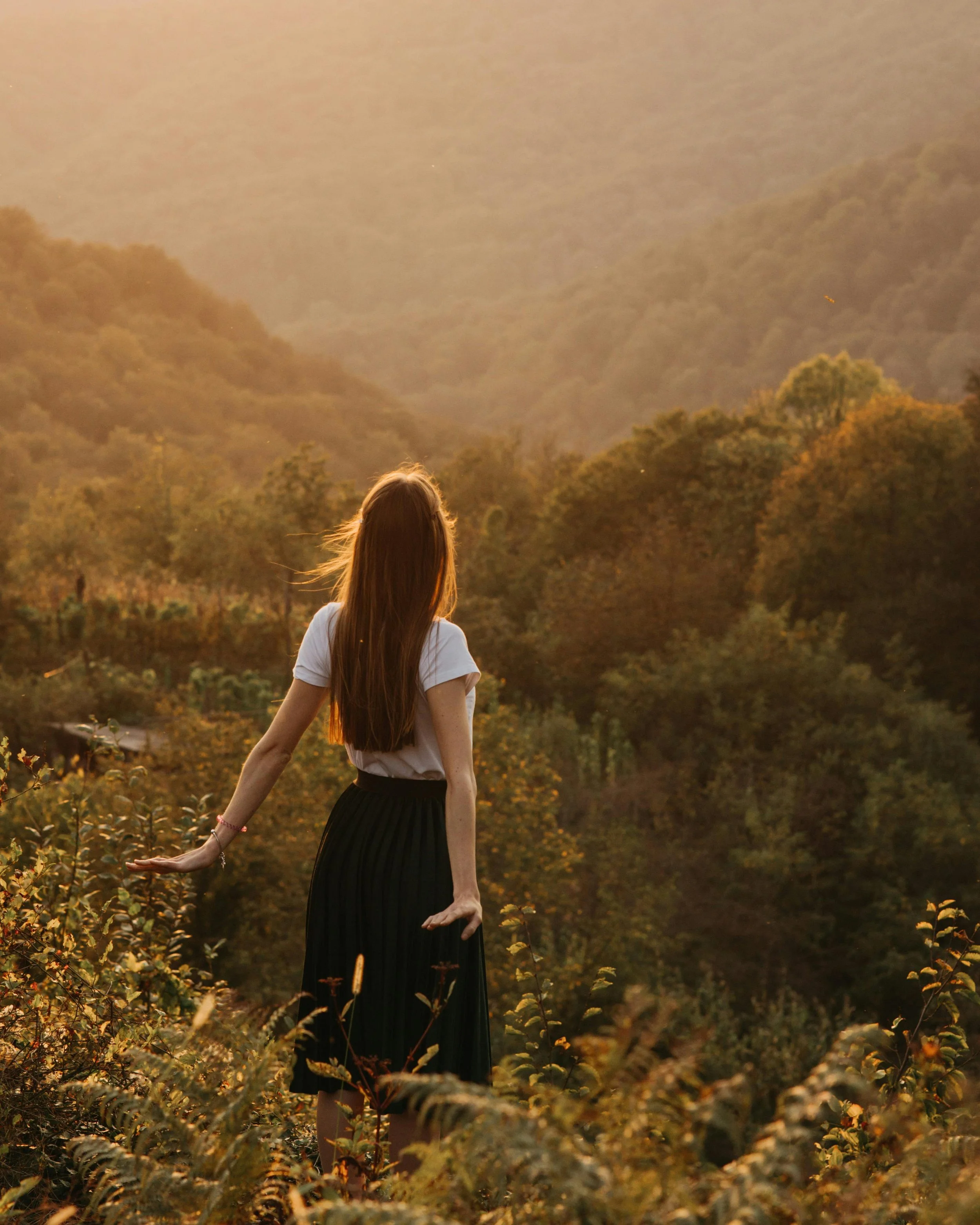 A woman with long hair and a white top standing in a field of plants at sunset, with mountainous terrain in the background.