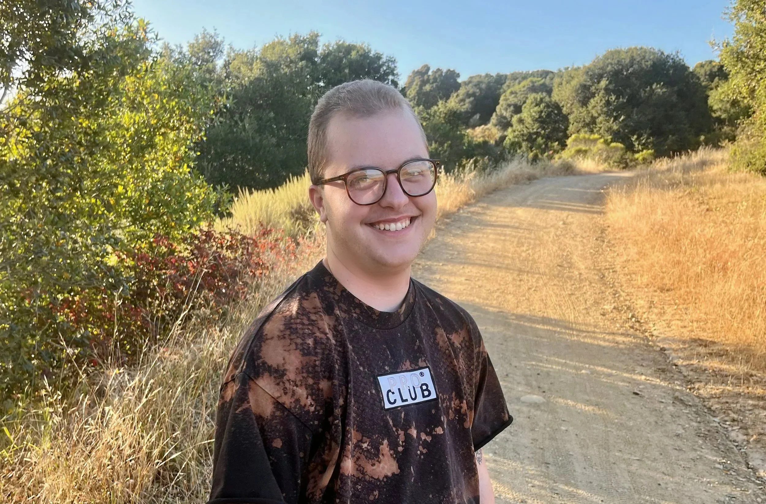 portrait of Elliott, founder and navigator at Radical Care Navigation. White queer with a buzzcut and glasses, smiling at the camera in the sun on a nature trail.