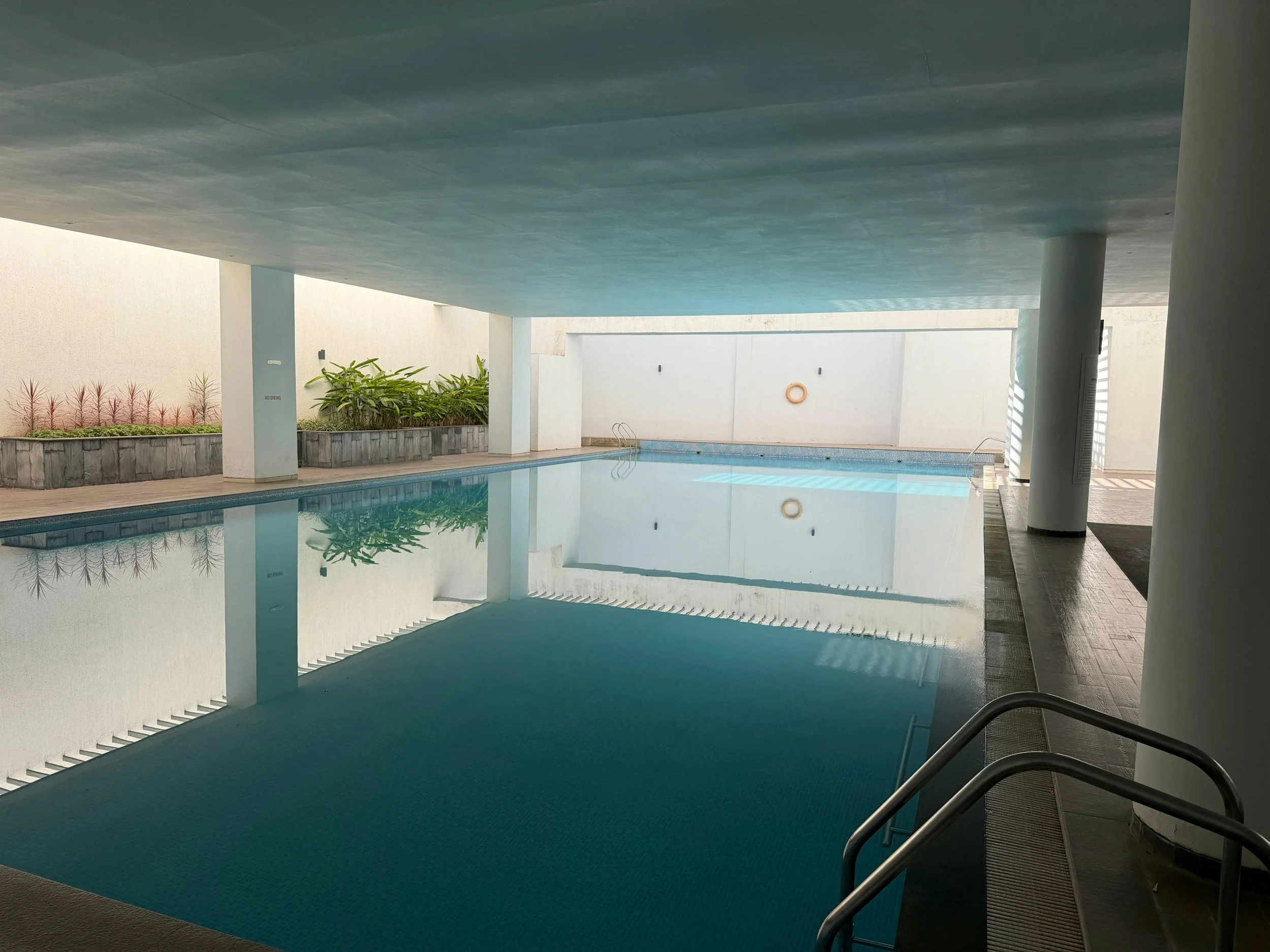 Indoor swimming pool with blue water, surrounded by white walls and covered ceiling, with stairs at the bottom right corner.
