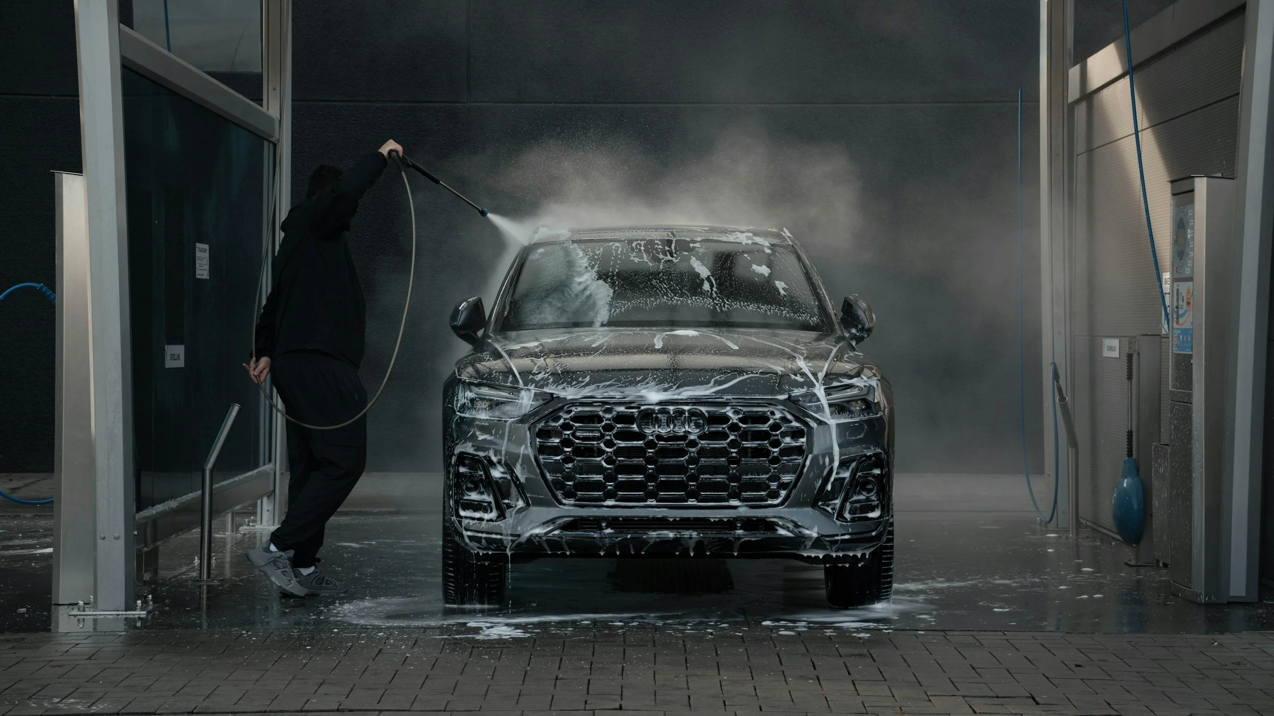 Person washing a silver Audi vehicle at a car wash station with soap and water.