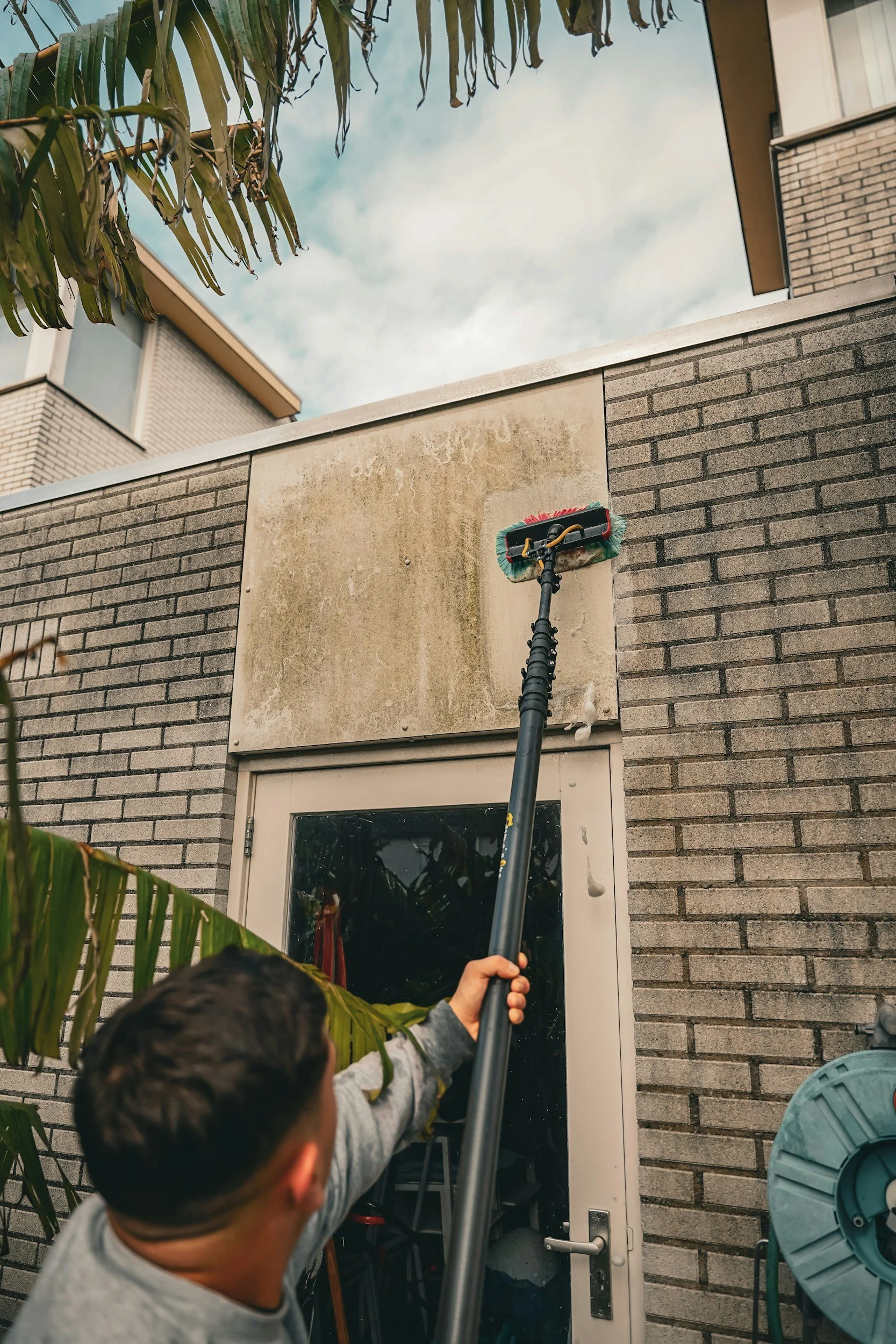 A child with short dark hair and gray shirt cleaning a window with a long-handled squeegee outside a brick building on a cloudy day.