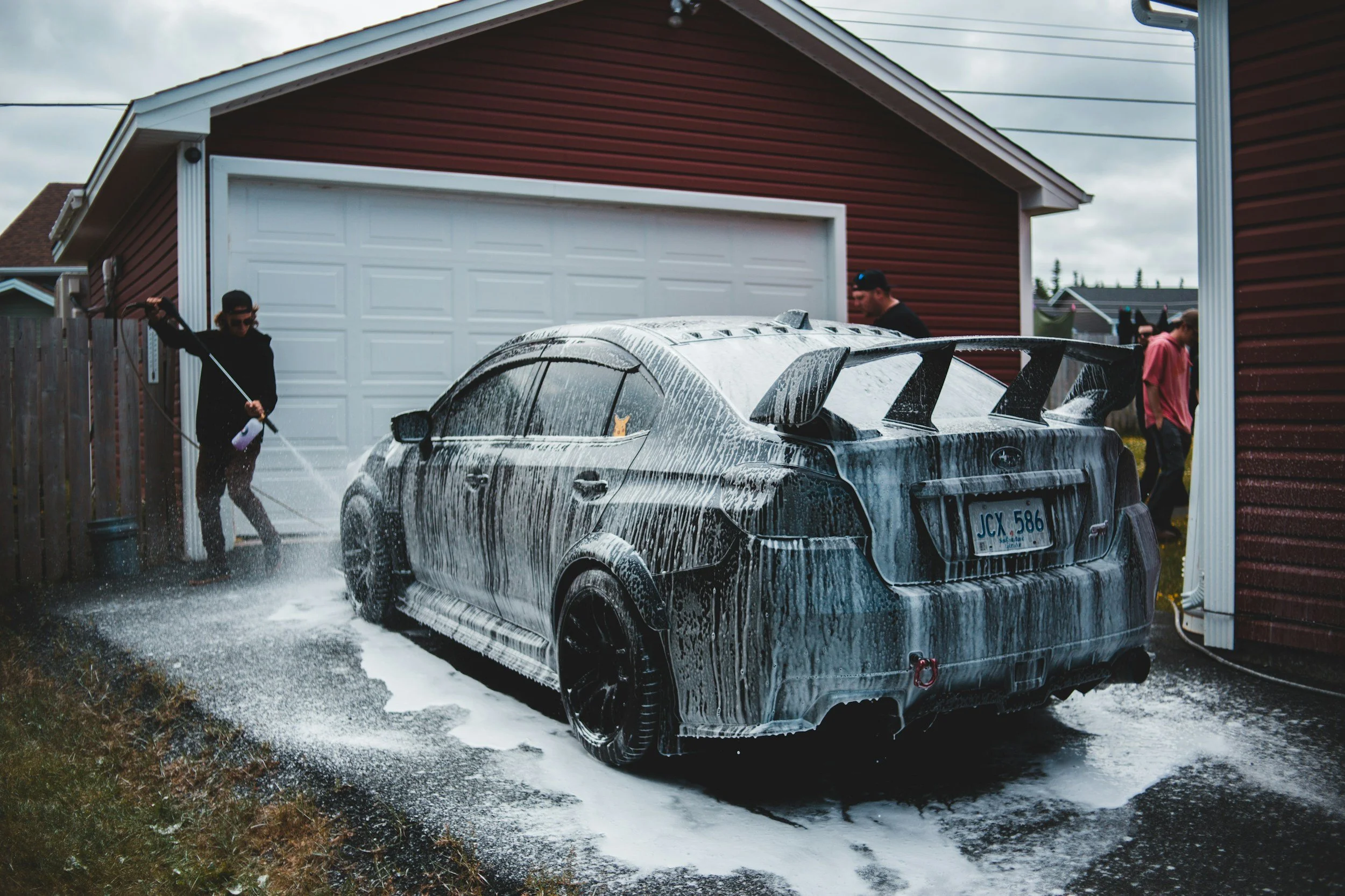 A black sports car being washed with soap suds in a residential driveway, with three people cleaning it.
