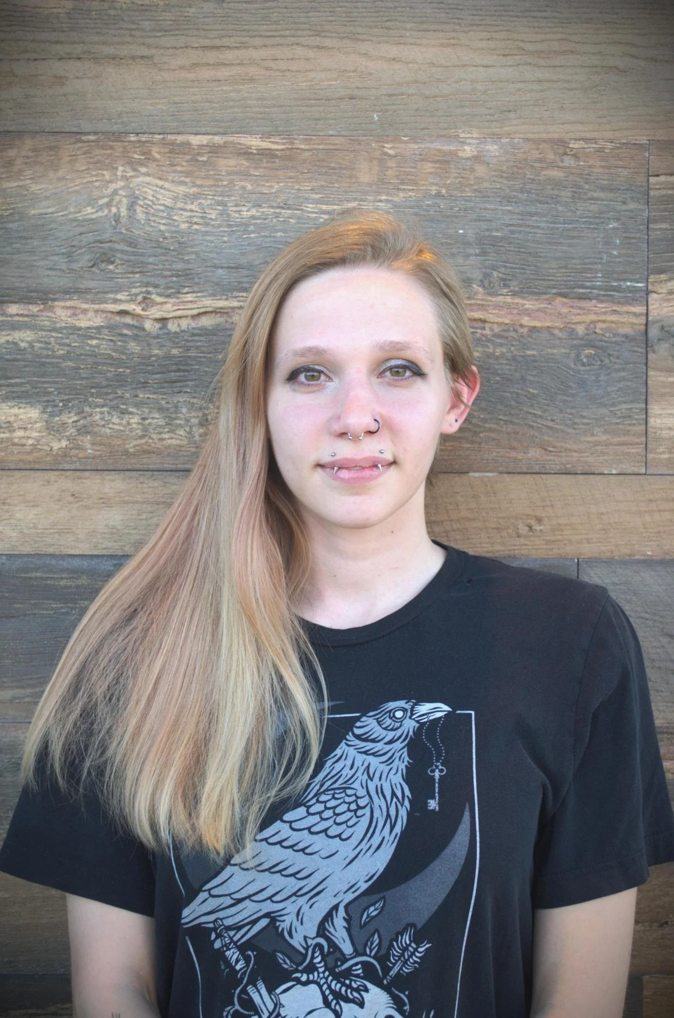 A young woman with long blonde hair, multiple facial piercings, and a nose ring, standing against a wooden wall, wearing a black graphic T-shirt with a detailed bird design.