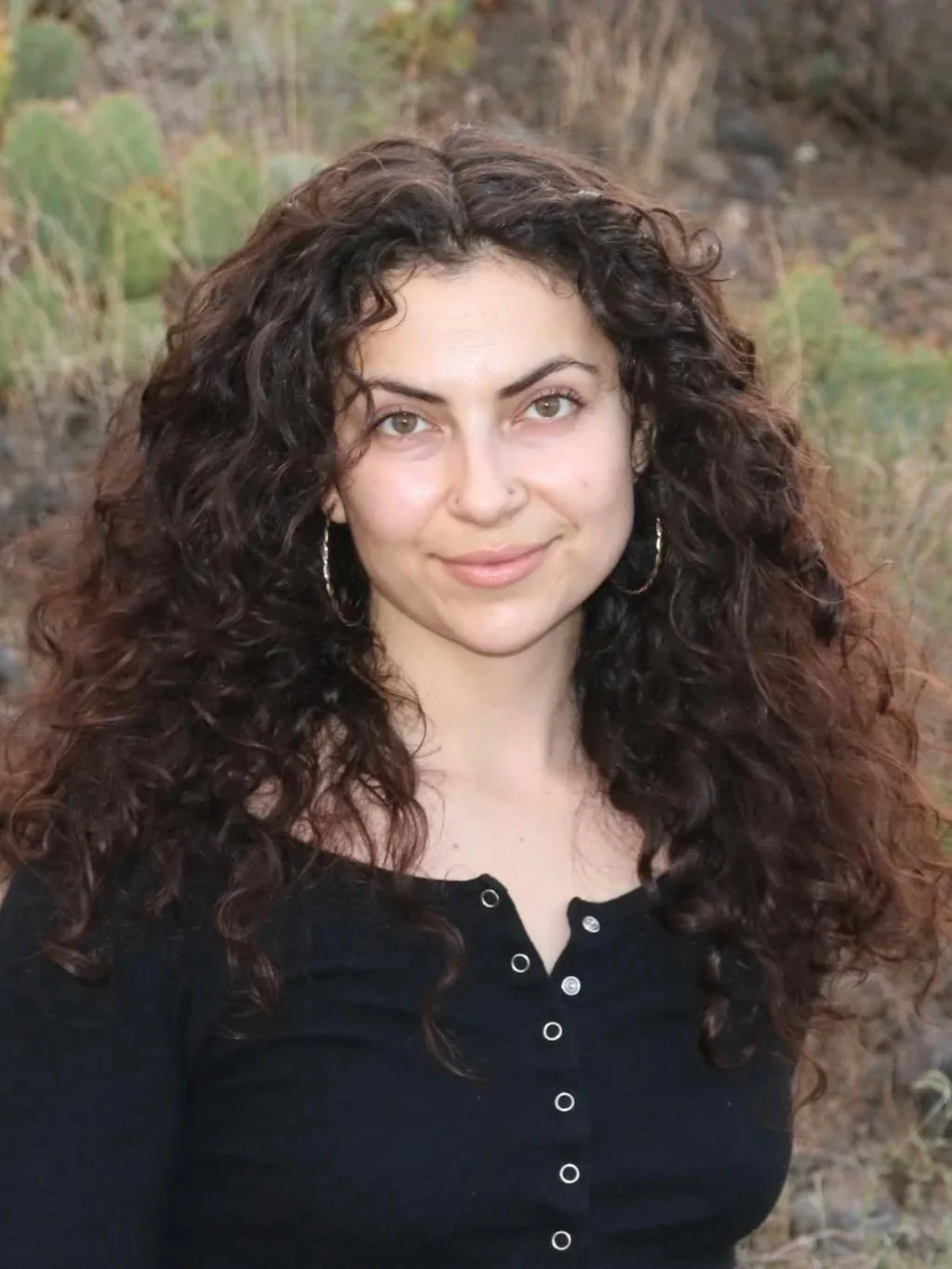 A woman with curly brown hair wearing a black top with buttons and hoop earrings, outdoors in a natural setting with green plants and rocky terrain in the background.