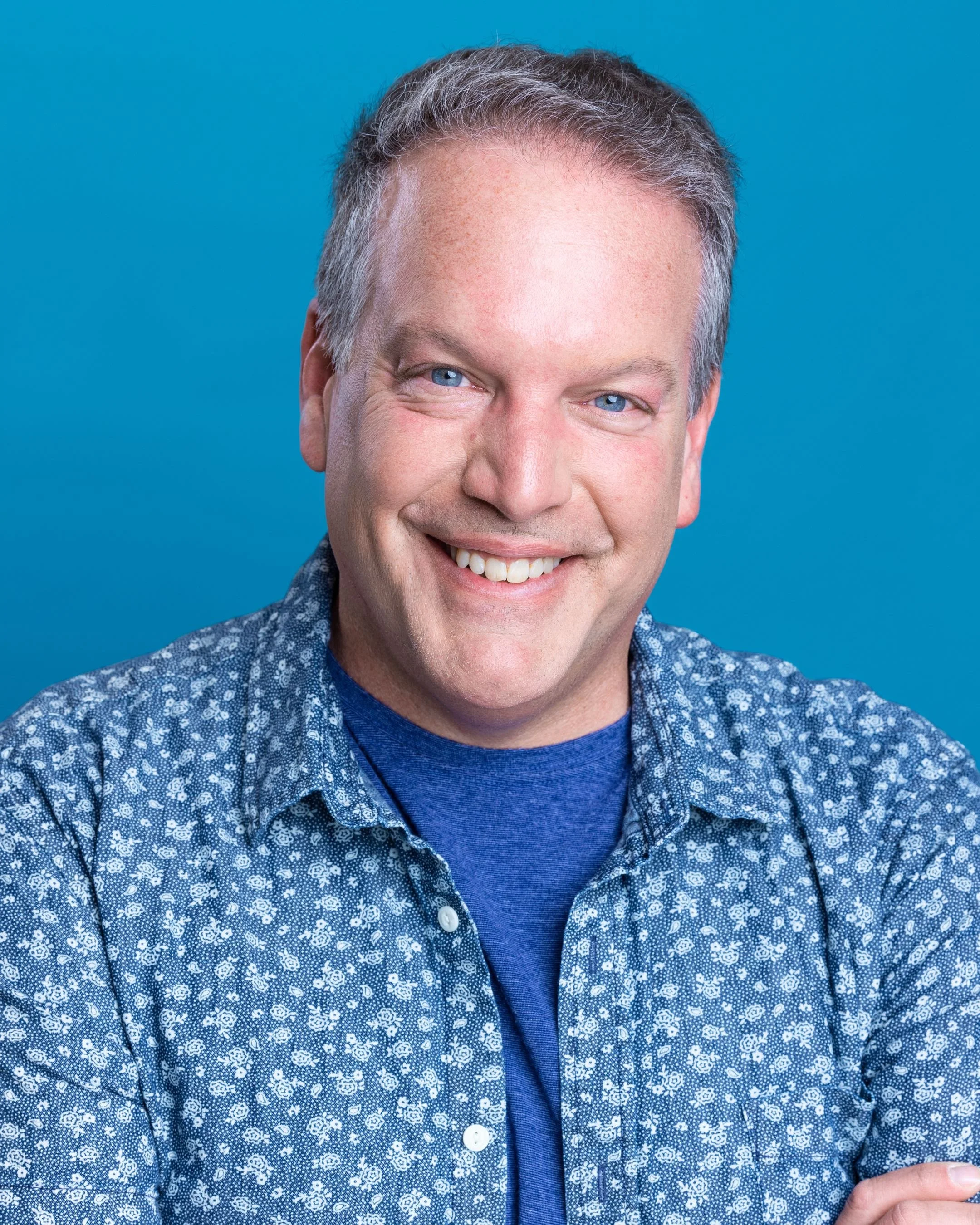 A smiling man with short gray hair, blue eyes, wearing a blue shirt and a patterned jacket, against a blue background.