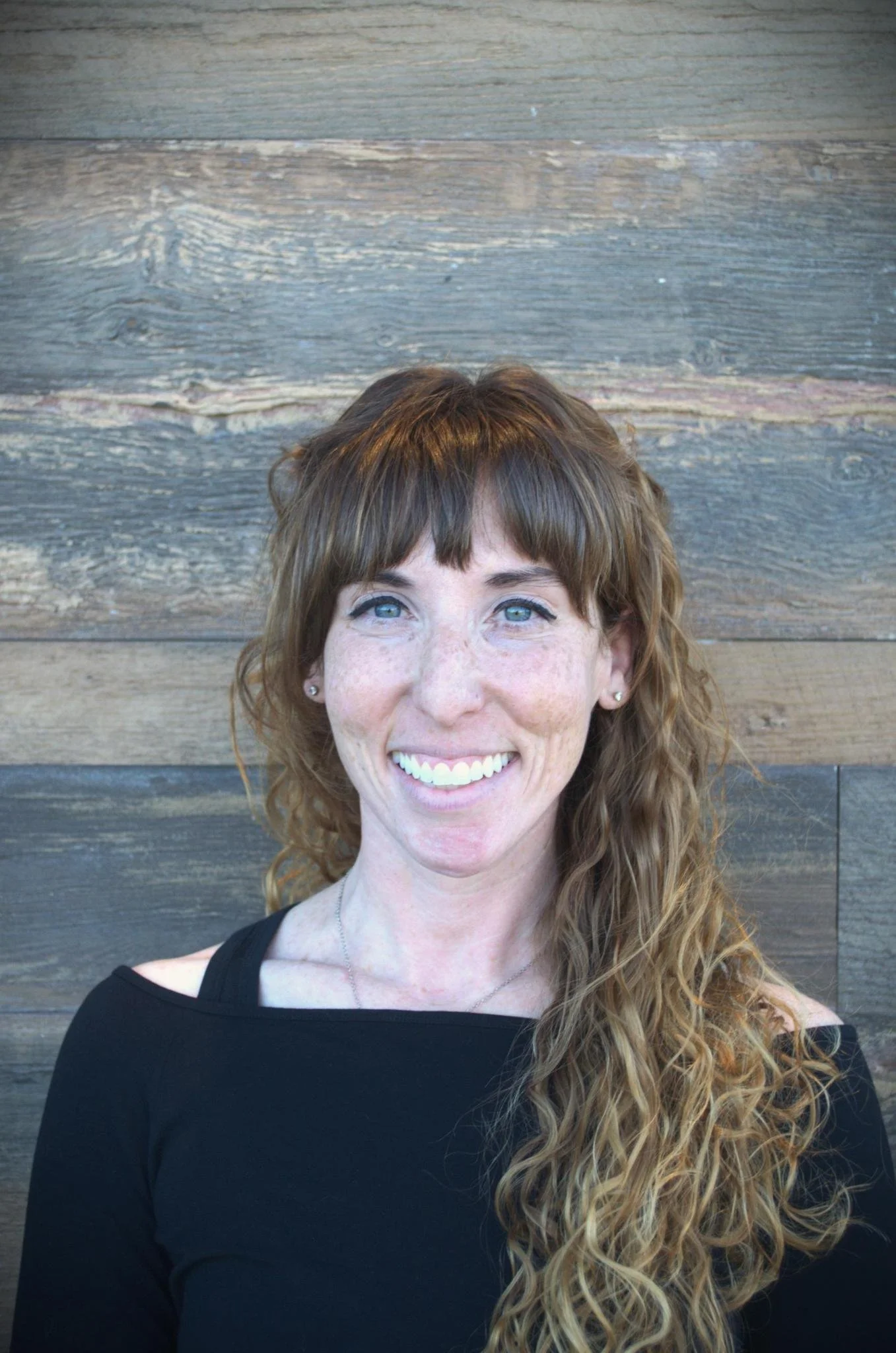 Close-up photo of a woman with long, wavy red hair, smiling, against a wooden wall background.
