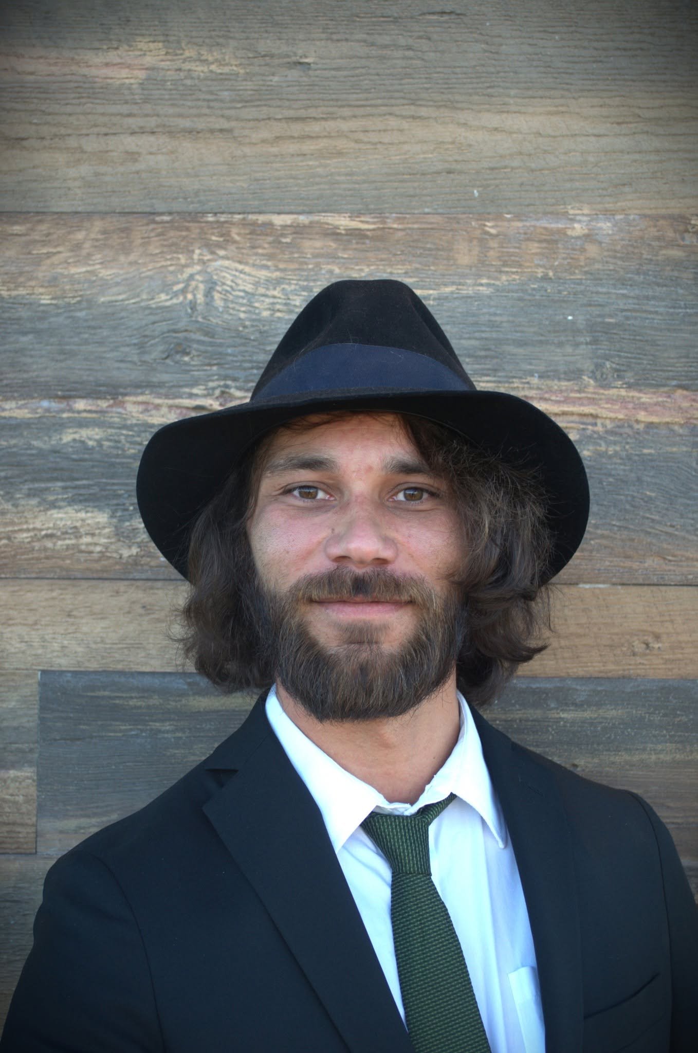 A man with long dark hair and a beard, dressed in a black suit, white shirt, and green tie, wearing a black hat, standing in front of a wooden background.