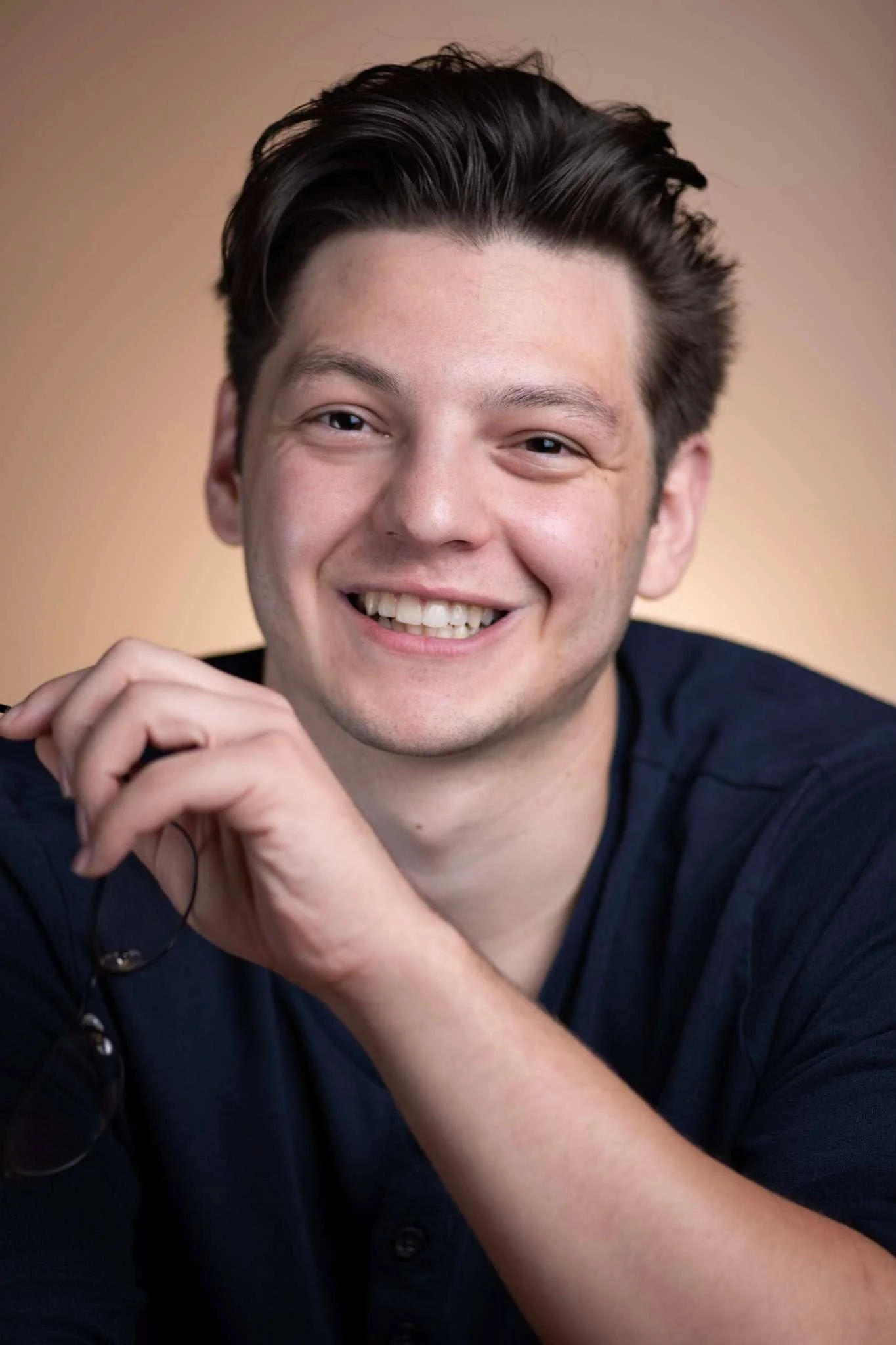Close-up portrait of a smiling man with dark hair, wearing a dark shirt, with hands clasped together.