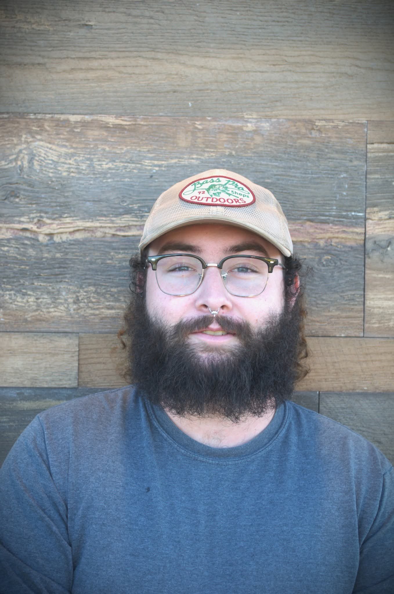 A man with a beard, glasses, and a nose ring wearing a beige cap with a Patch Pass logo, standing in front of a wooden wall.