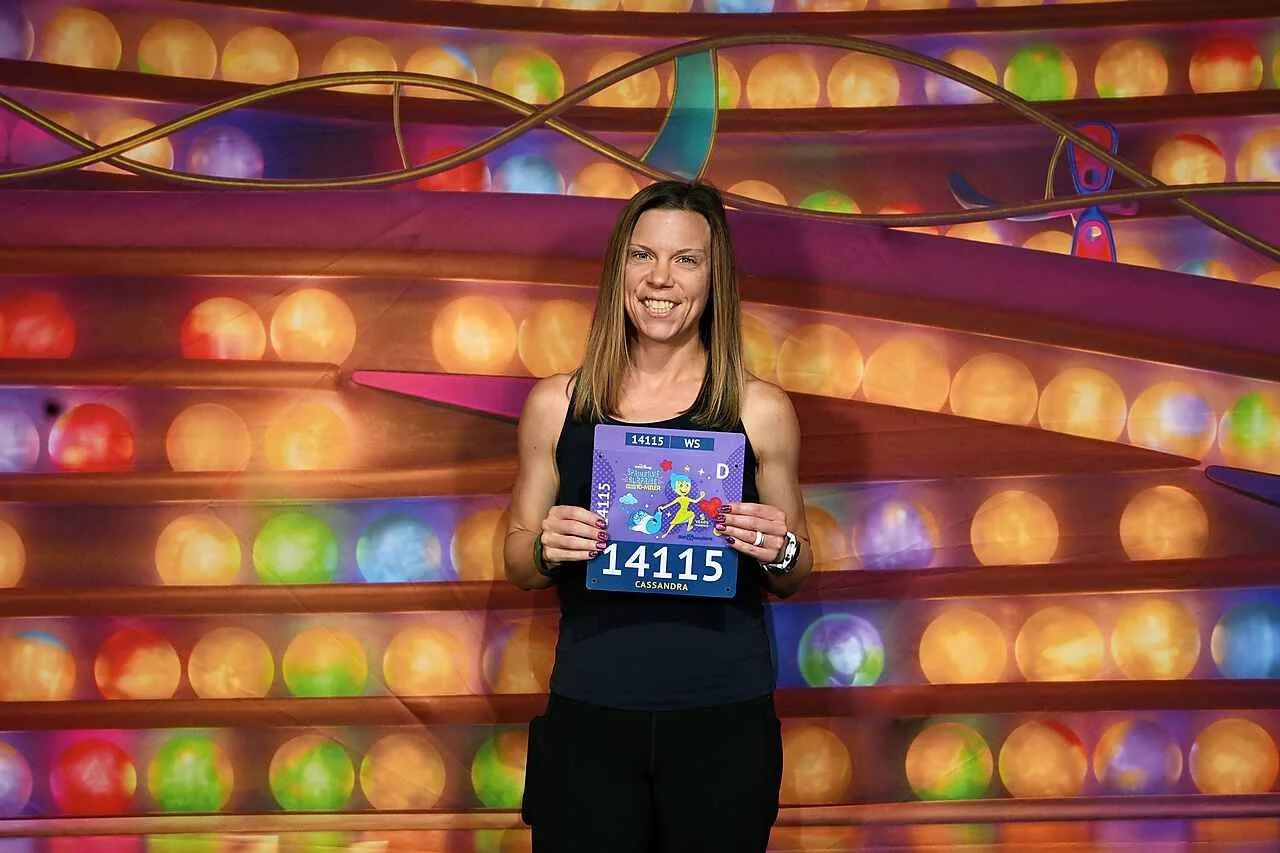Woman standing in front of a colorful, illuminated background, holding a purple sign with the number 14115 and a mermaid illustration, smiling at the camera.