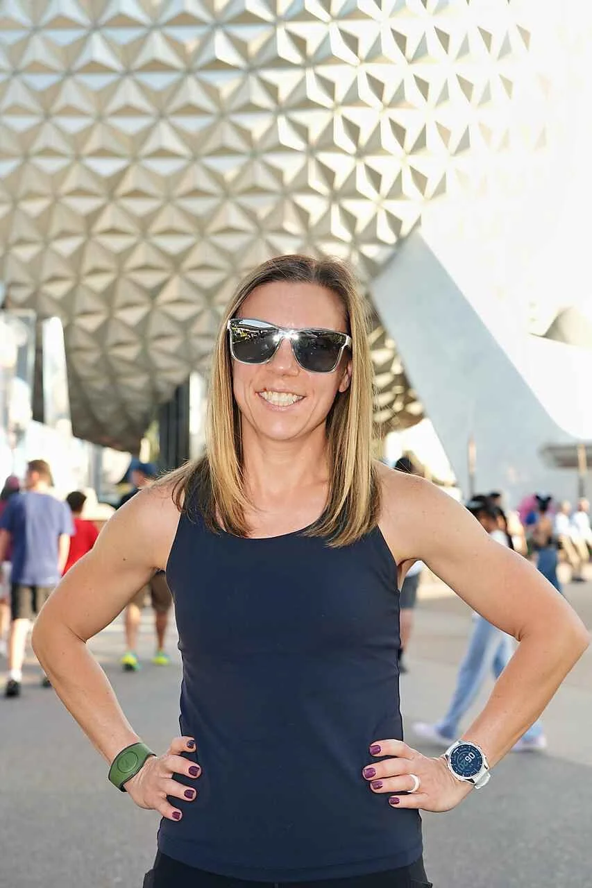 Smiling woman wearing sunglasses, a black tank top, a white watch, and a green fitness tracker, standing outdoors in front of the Spaceship Earth geodesic sphere at EPCOT in Disney World during daytime.