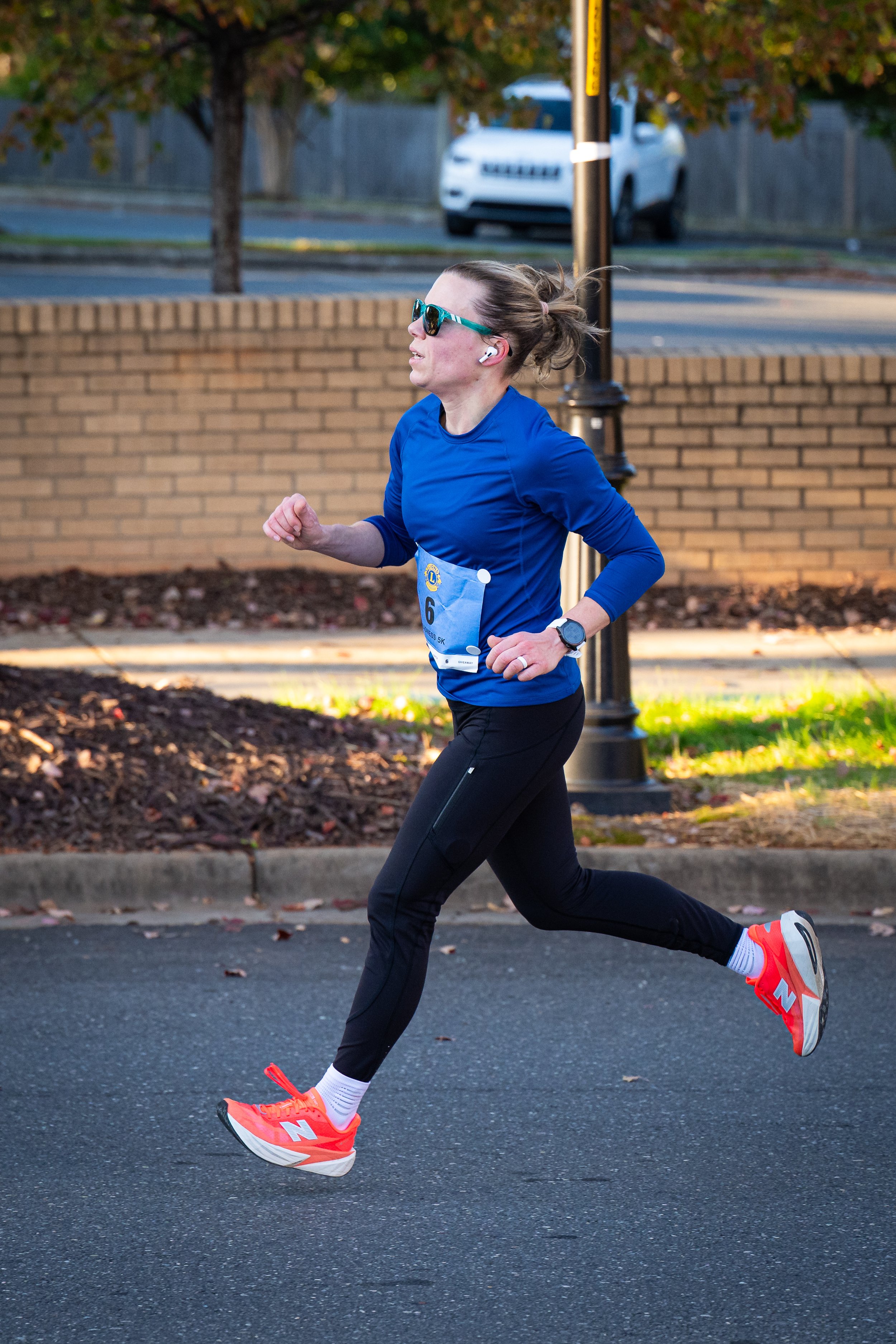 A woman running outdoors, wearing a blue long-sleeve shirt, black leggings, bright orange running shoes, sunglasses, and wireless earbuds.